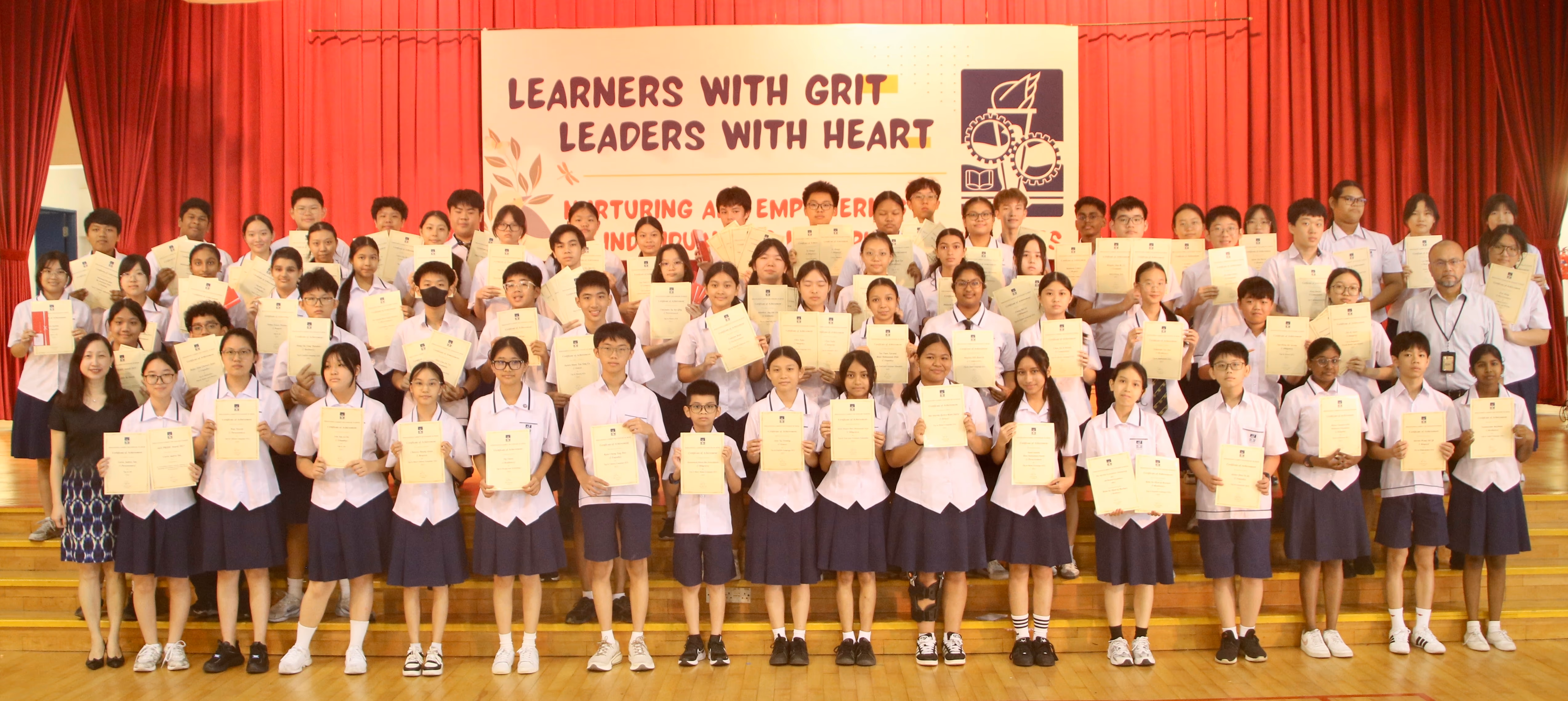 Large group of uniformed students holding certificates on a stage with a banner reading Learners With Grit Leaders With Heart.