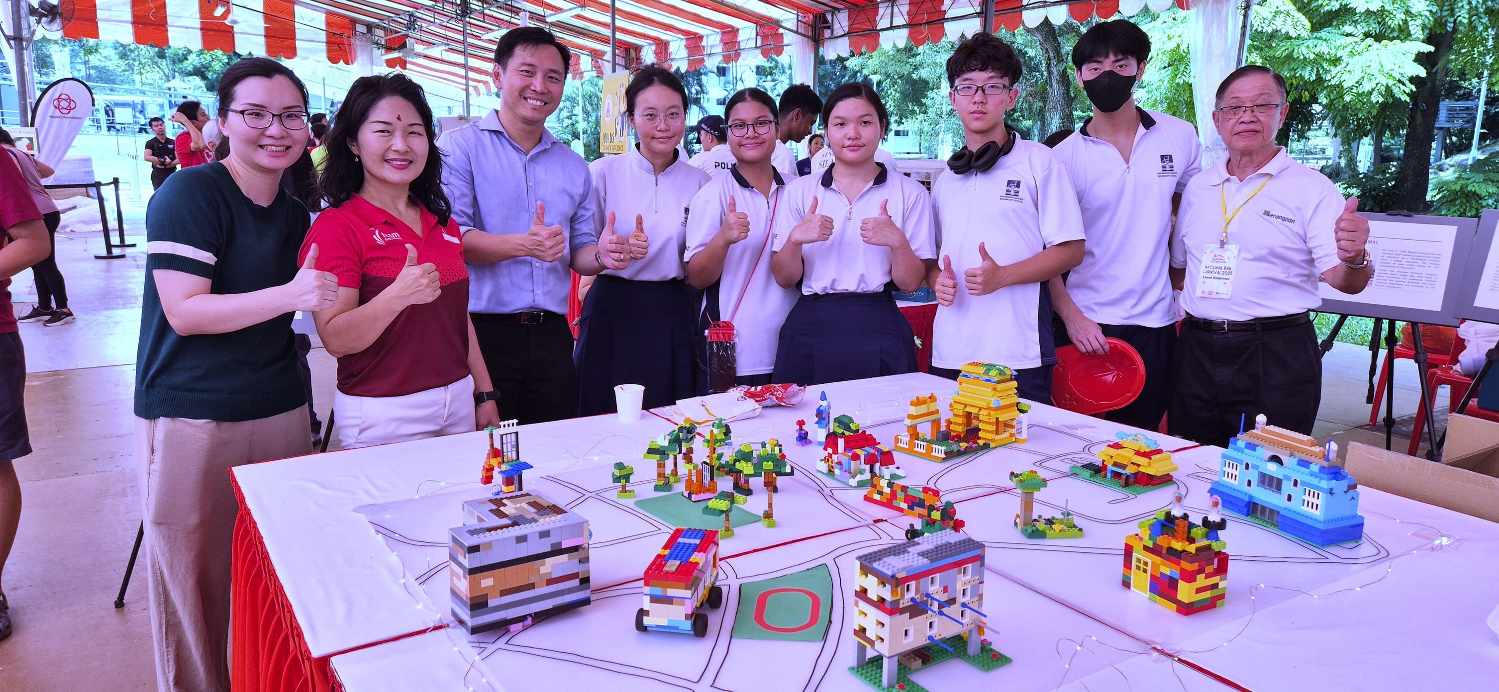 A group of nine people give thumbs up behind a table displaying colorful LEGO city models at an outdoor event.