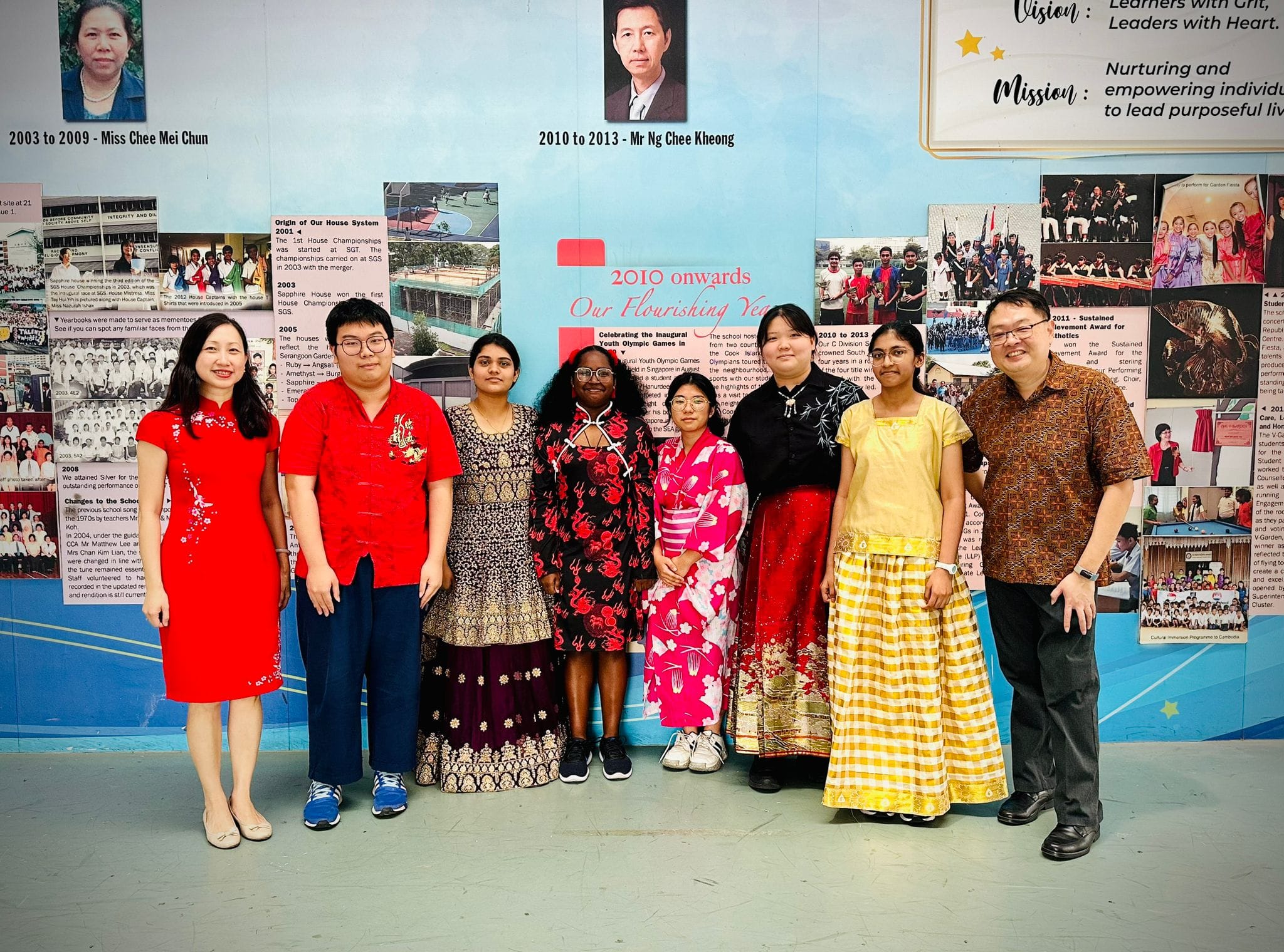 Eight people in colorful traditional outfits pose in front of a school history display wall.