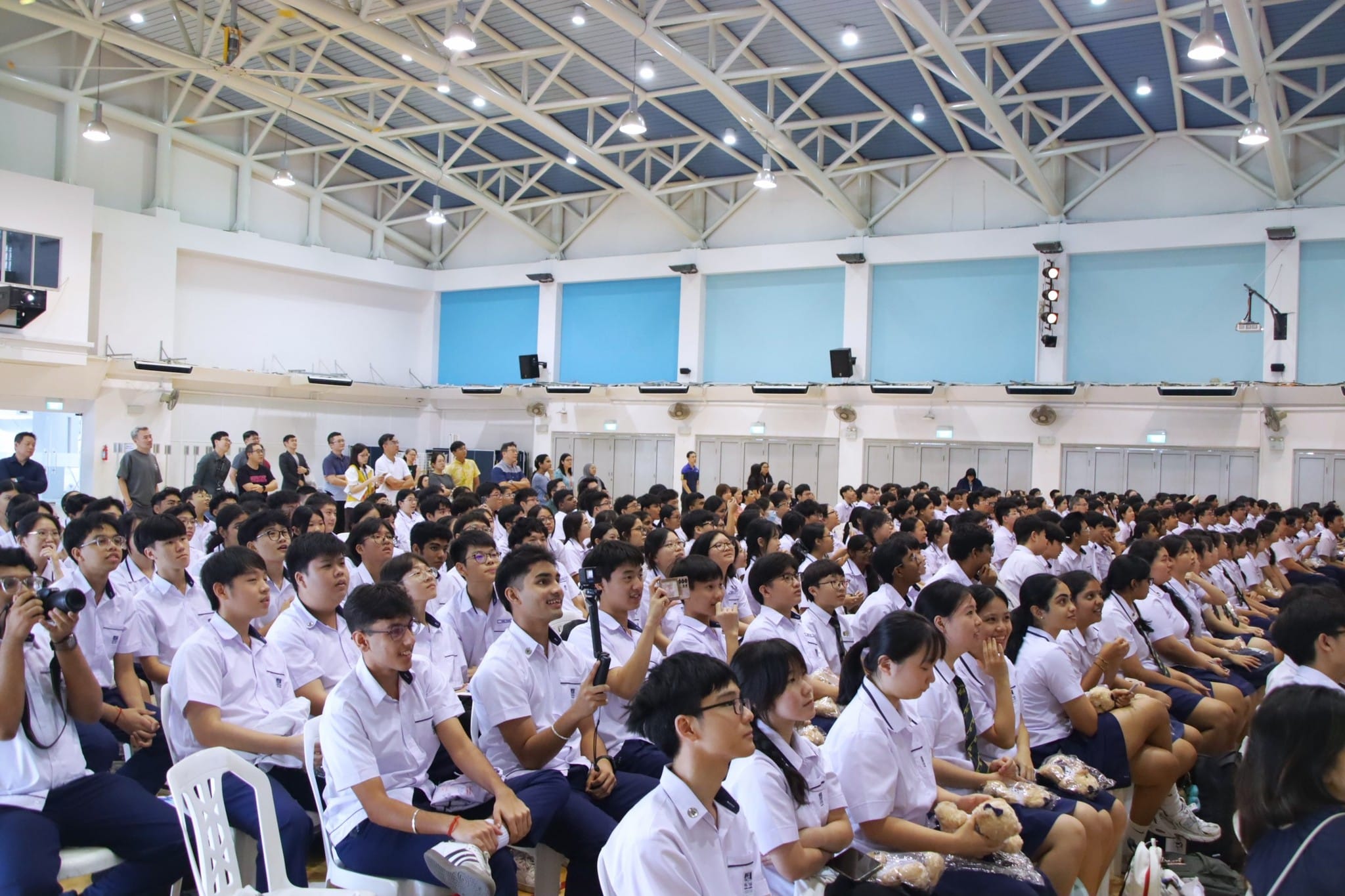 Large crowd of students in white uniforms seated in a school hall during an assembly event.