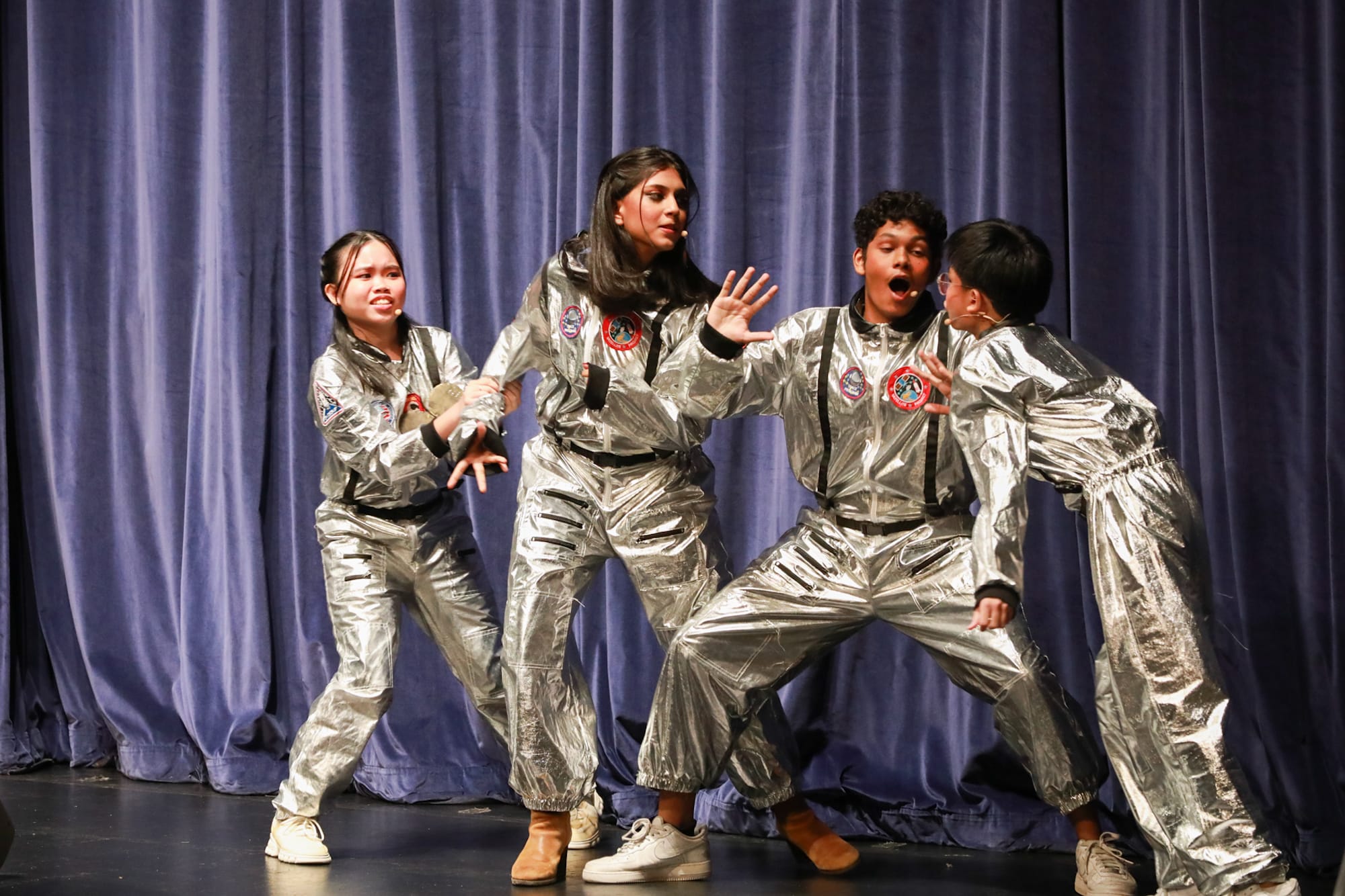 Four teens in silver astronaut jumpsuits perform on a stage with a blue curtain backdrop.