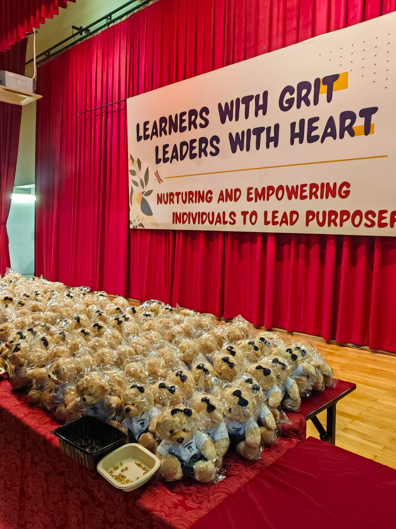 Rows of wrapped teddy bears on a red table before a stage banner reading Learners With Grit Leaders With Heart.