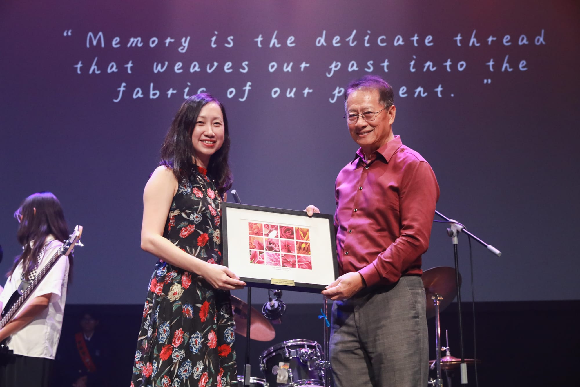 Two people smile while holding a framed floral photo collage on stage, with a quote projected behind them.