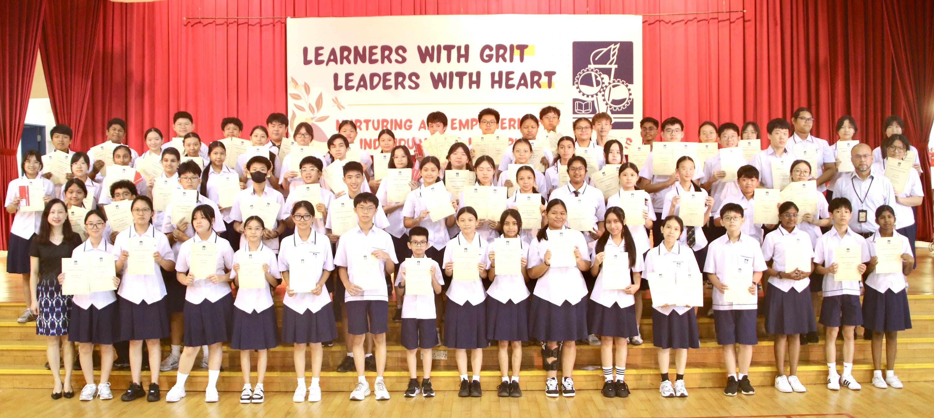 Large group of uniformed school students holding certificates on a stage with a banner reading Learners With Grit Leaders With Heart.