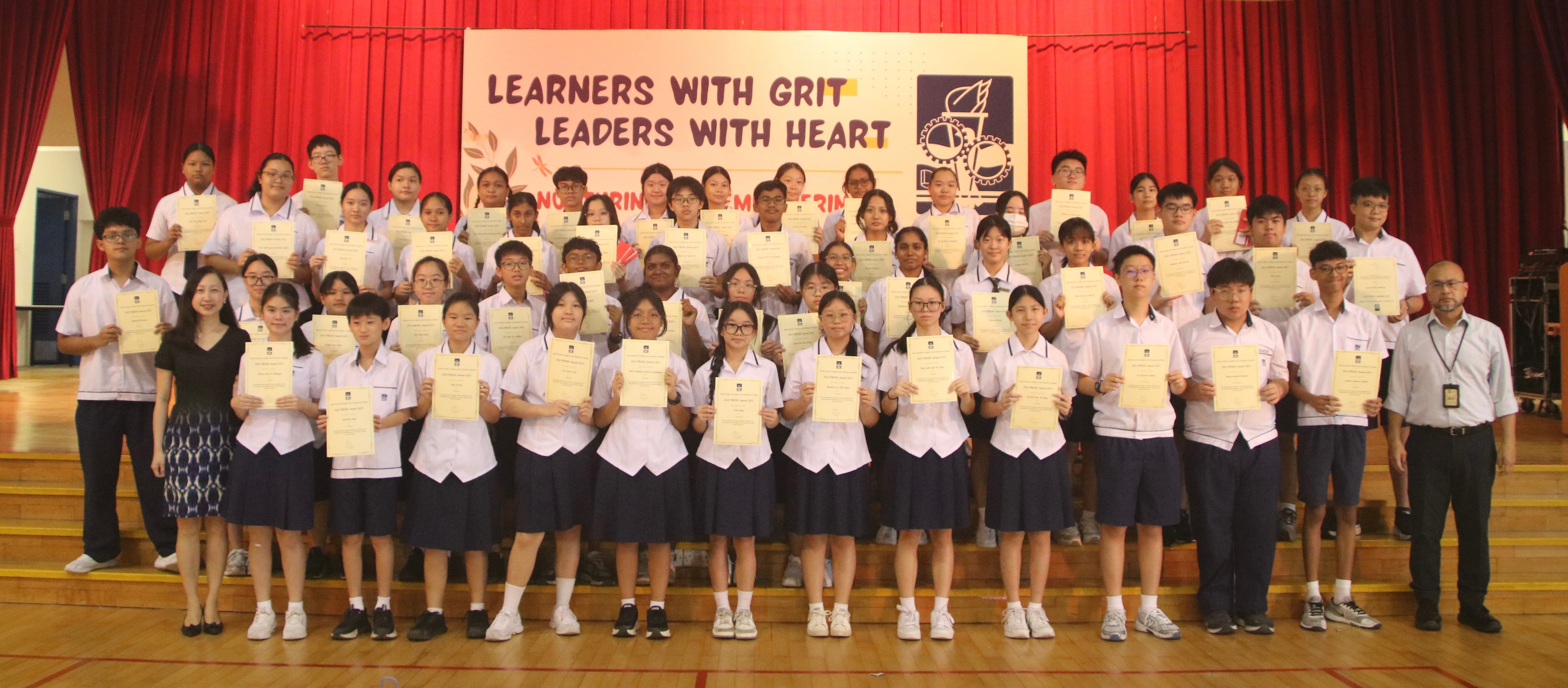Students in school uniforms hold certificates on a stage before a banner reading Learners With Grit Leaders With Heart.