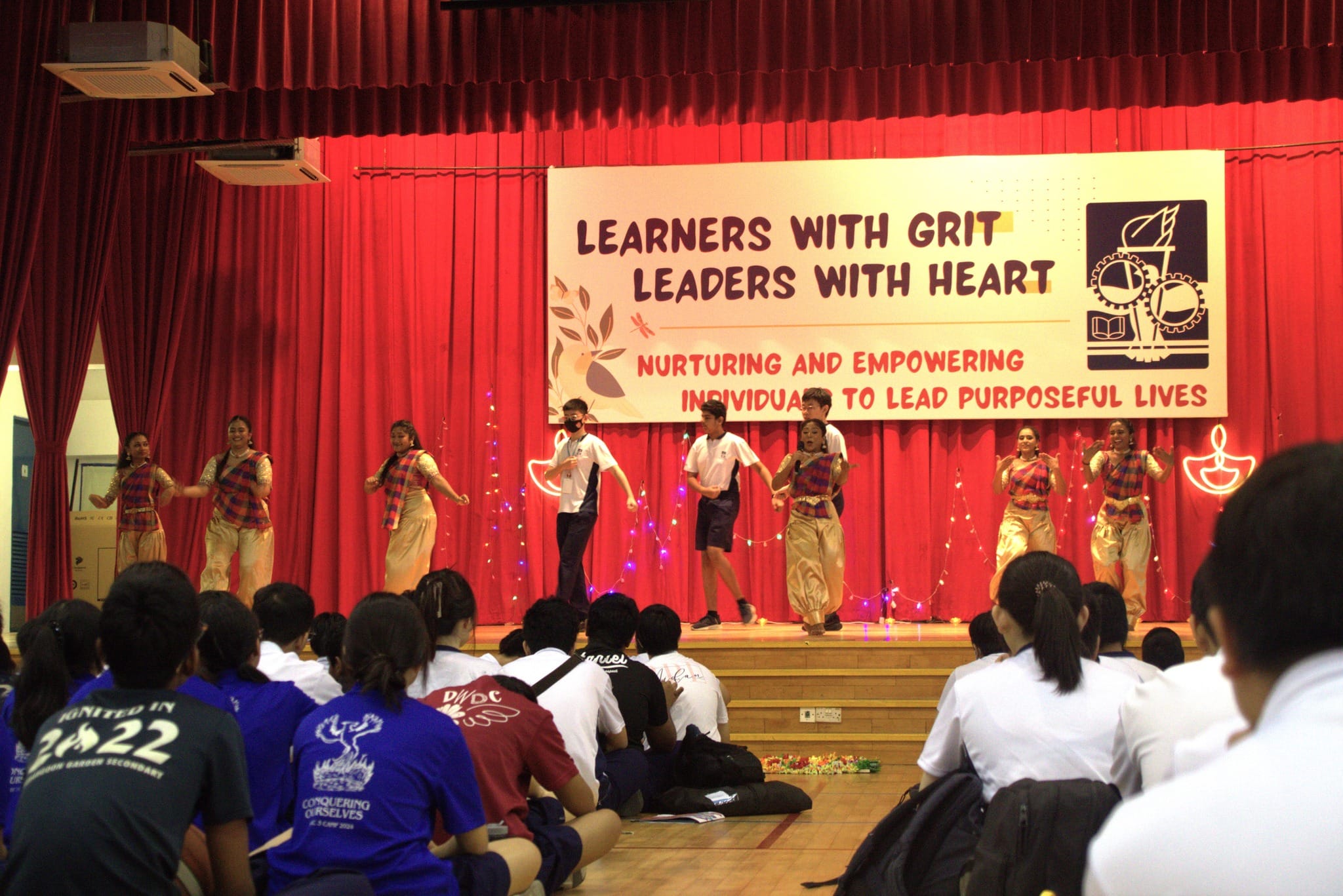 Students perform a cultural dance on stage before seated audience under a banner reading Learners With Grit Leaders With Heart.