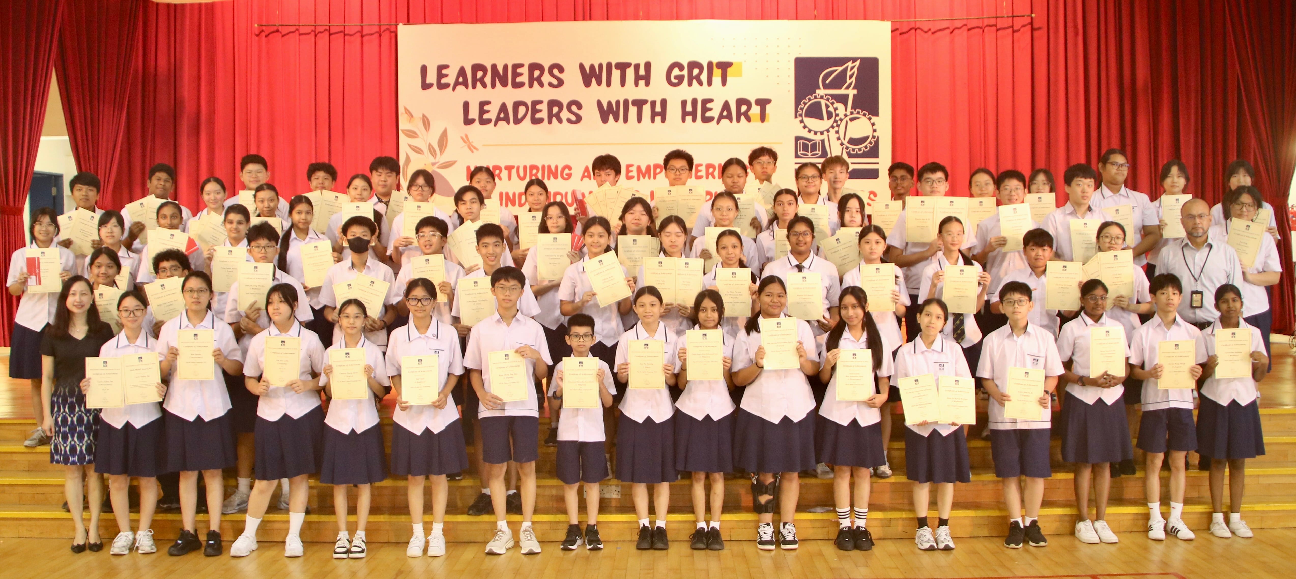 Large group of uniformed students holding certificates on a stage with a banner reading Learners With Grit Leaders With Heart.