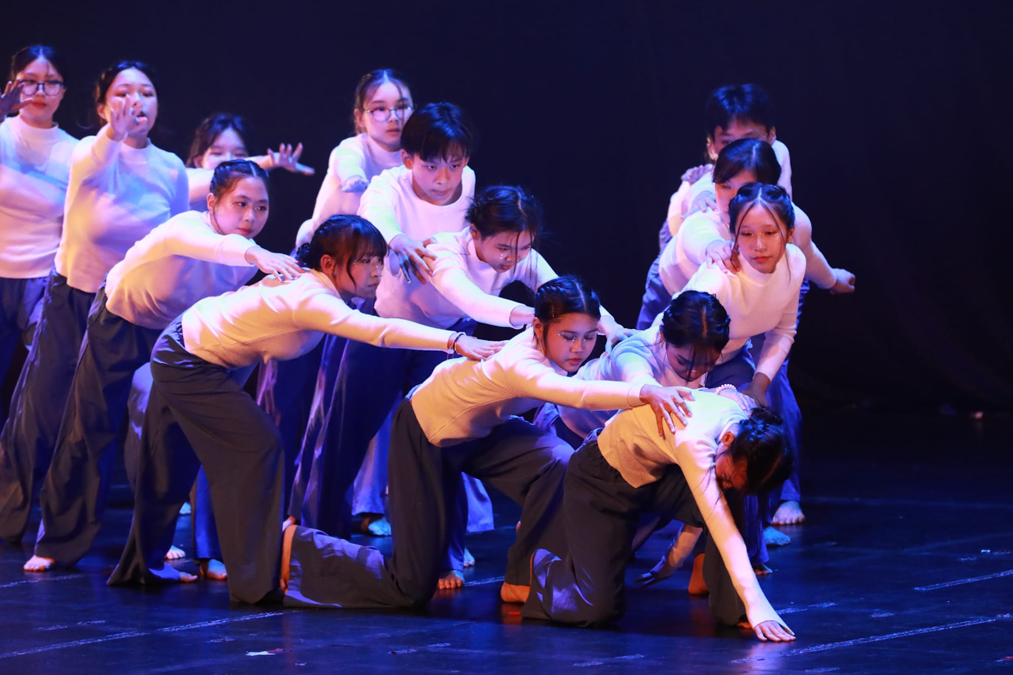 A group of young dancers in white tops and dark pants perform a dramatic routine on a dimly lit stage.