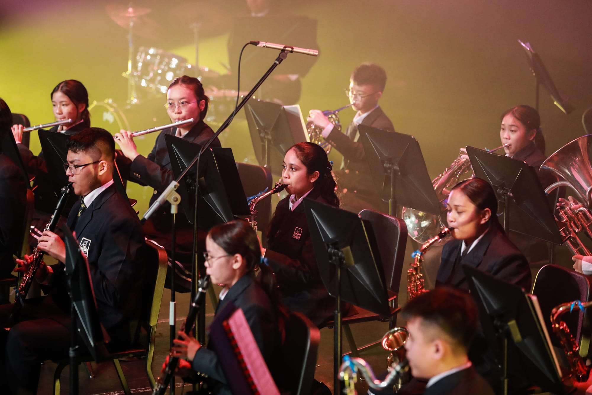 Students in dark uniforms perform as a wind band on a lit stage, playing flutes, clarinets, saxophones, and brass instruments.