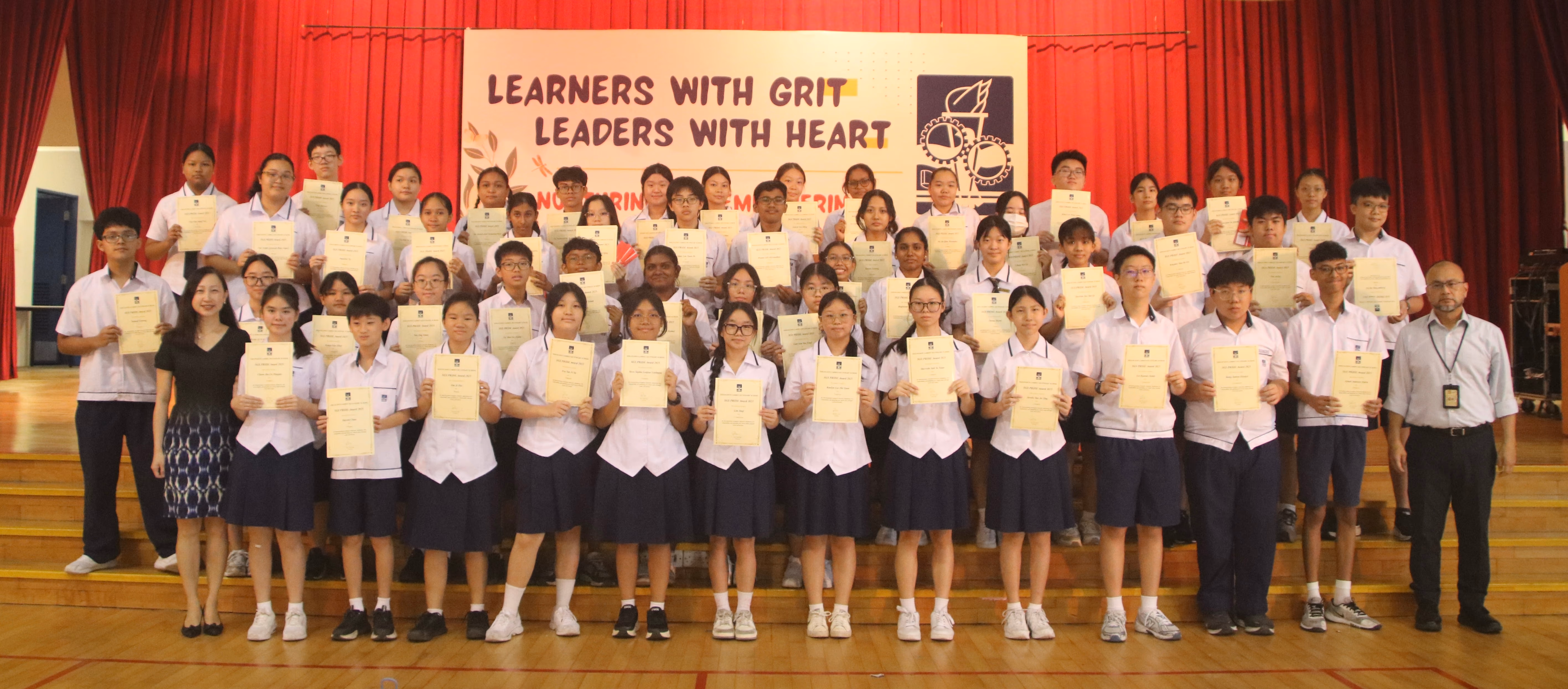 Students in school uniforms hold certificates on a stage before a banner reading Learners With Grit Leaders With Heart.