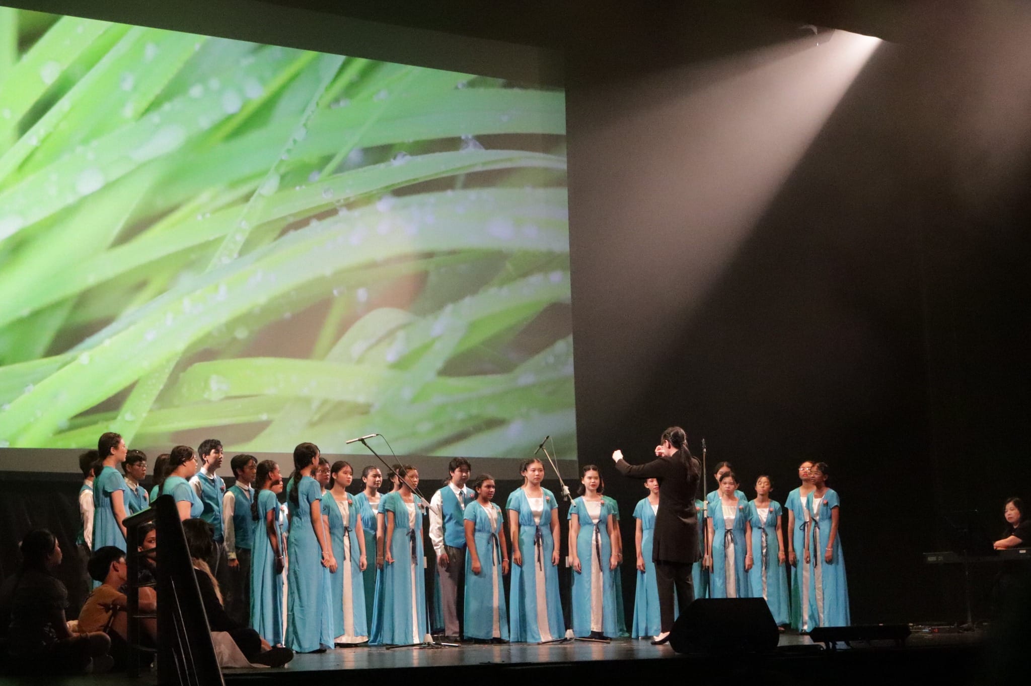 A choir in teal dresses performs on a dark stage with a conductor, lit by a spotlight, before a green leaf projection.