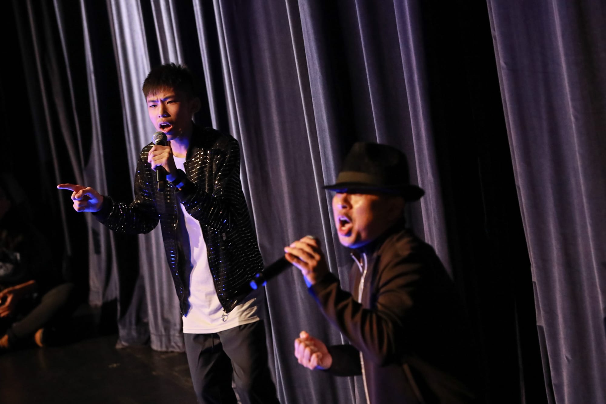 Two men singing into microphones on a dark stage in front of a gray curtain, one wearing a sequined jacket.