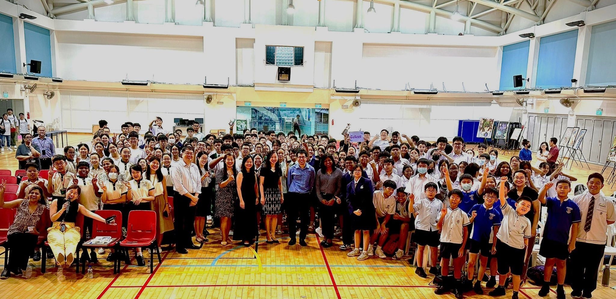 Large group of students and teachers posing together in a school gymnasium with wooden floors and high ceilings.