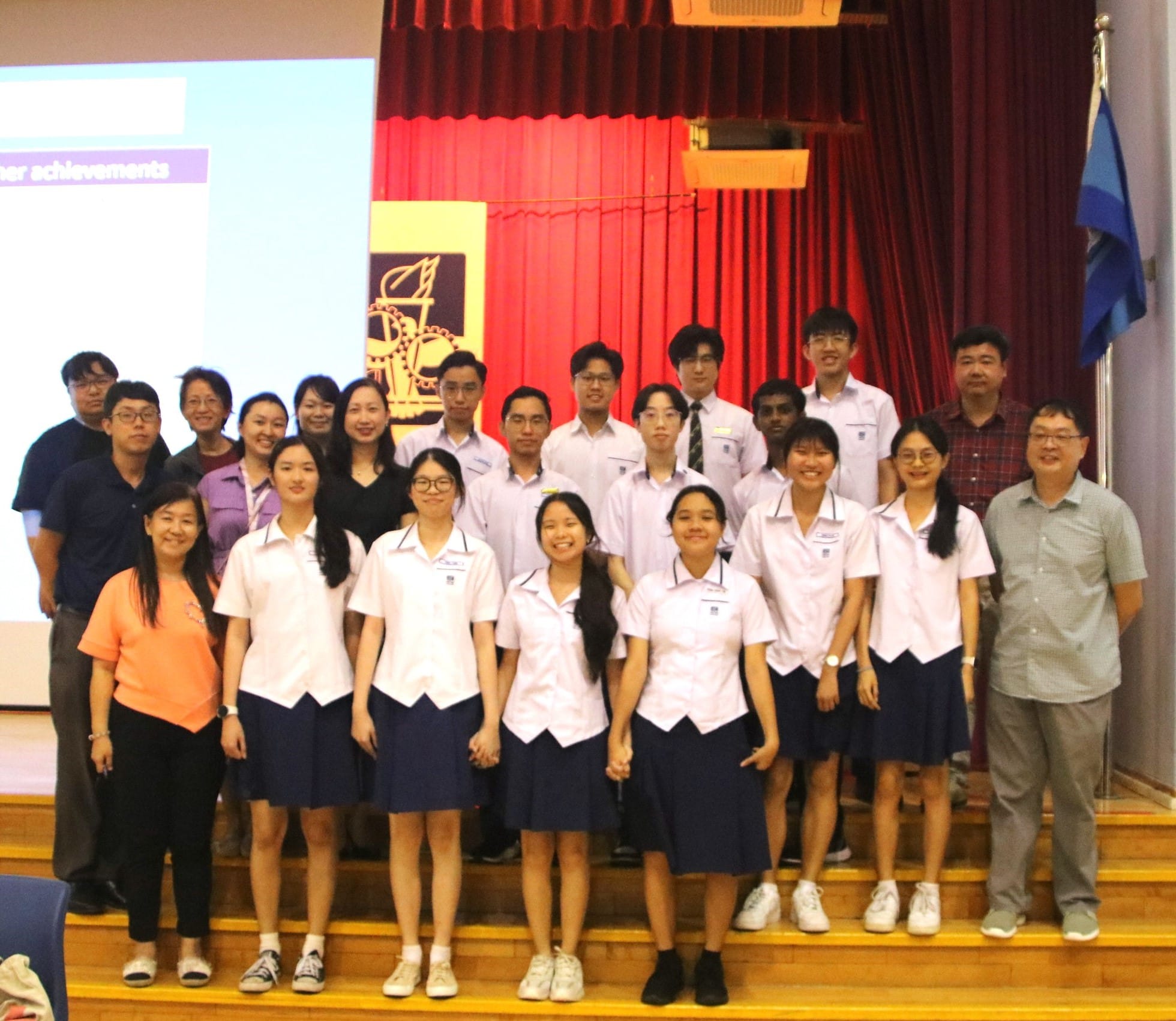 A group of students in white and navy uniforms pose with adults on a school stage with red curtains behind them.