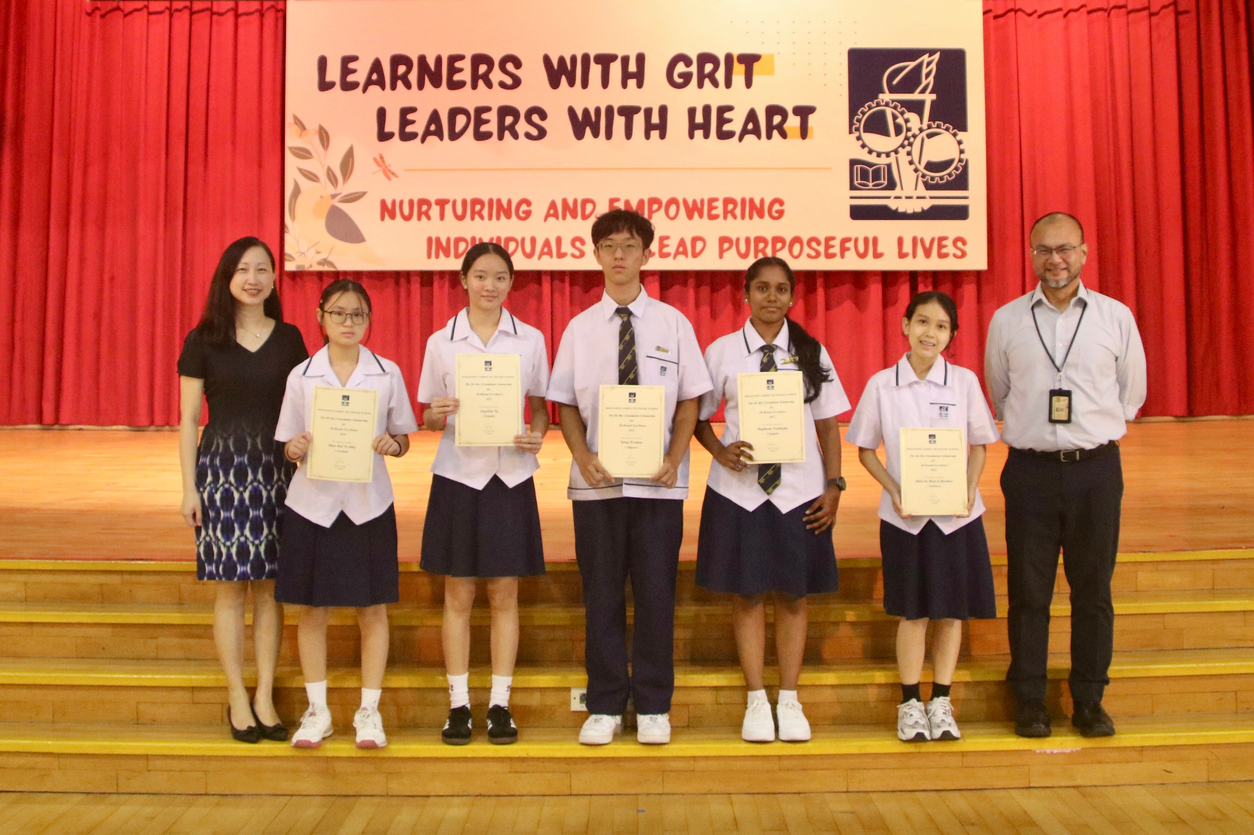 Four students in uniforms hold certificates on a stage, flanked by two adults, in front of a school banner.