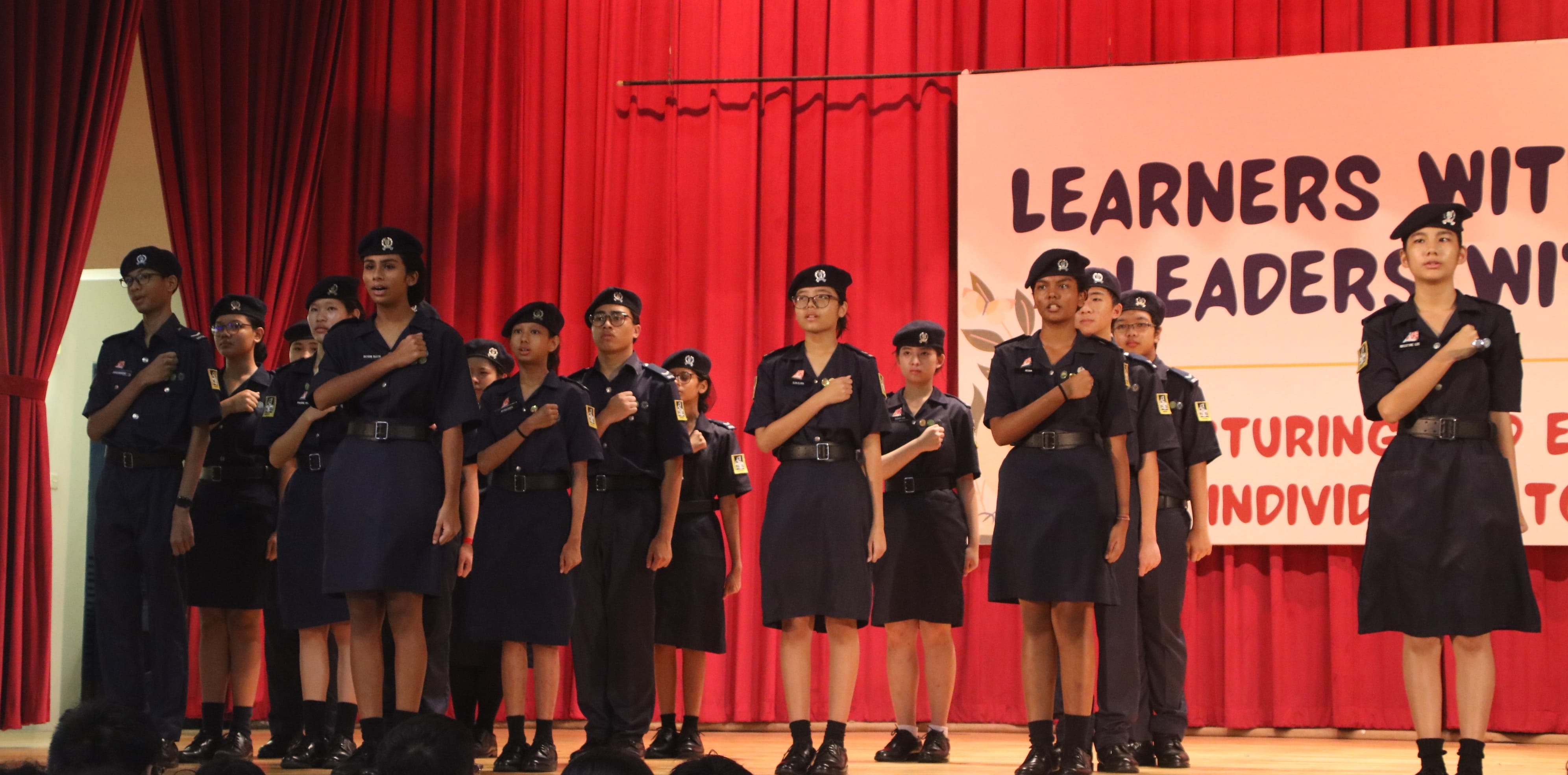 Photograph of a group of uniformed students standing on stage in formation with hands over their chests, likely during a pledge or anthem. Red curtain backdrop and a partially visible sign with motivational text emphasize a formal school or leadership event.