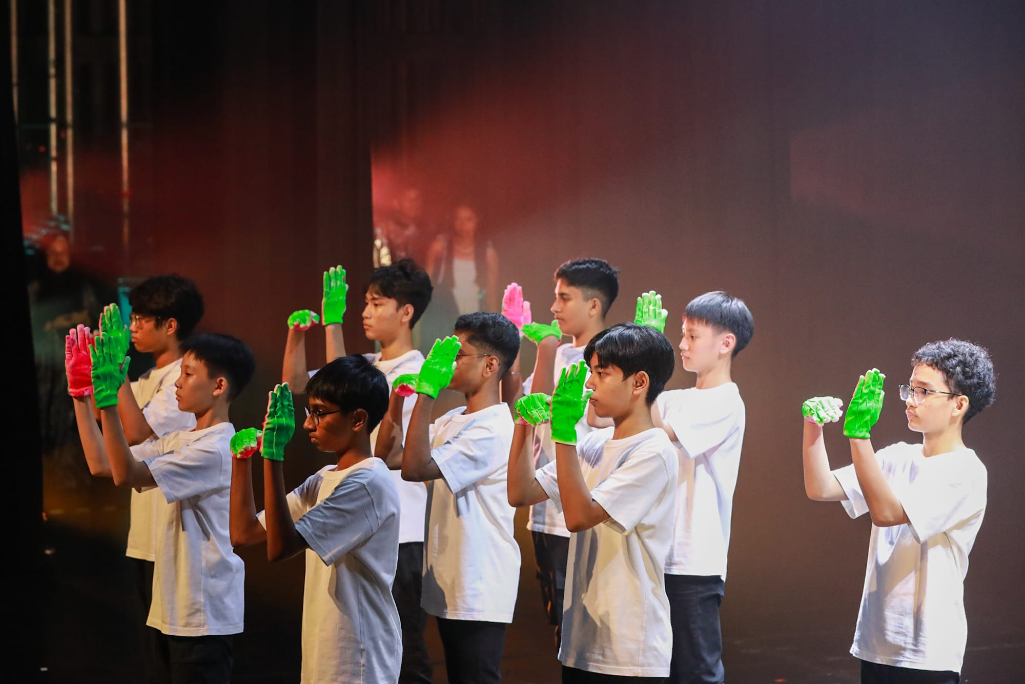 Boys in white t-shirts stand in rows on a dark stage, raising hands wearing bright green and pink gloves.