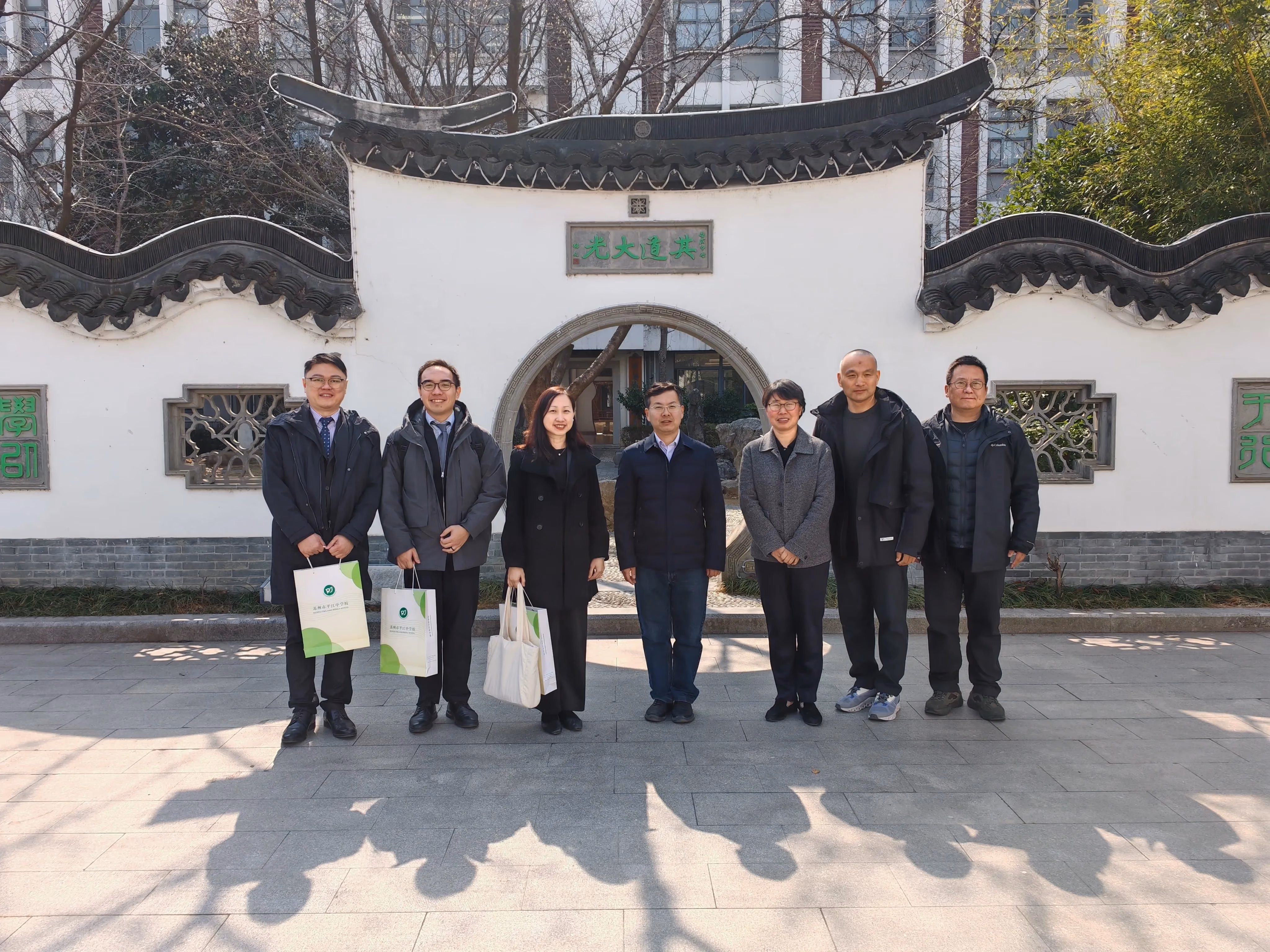 Seven people pose in front of a white traditional Chinese gate arch with green characters and curved roof tiles.