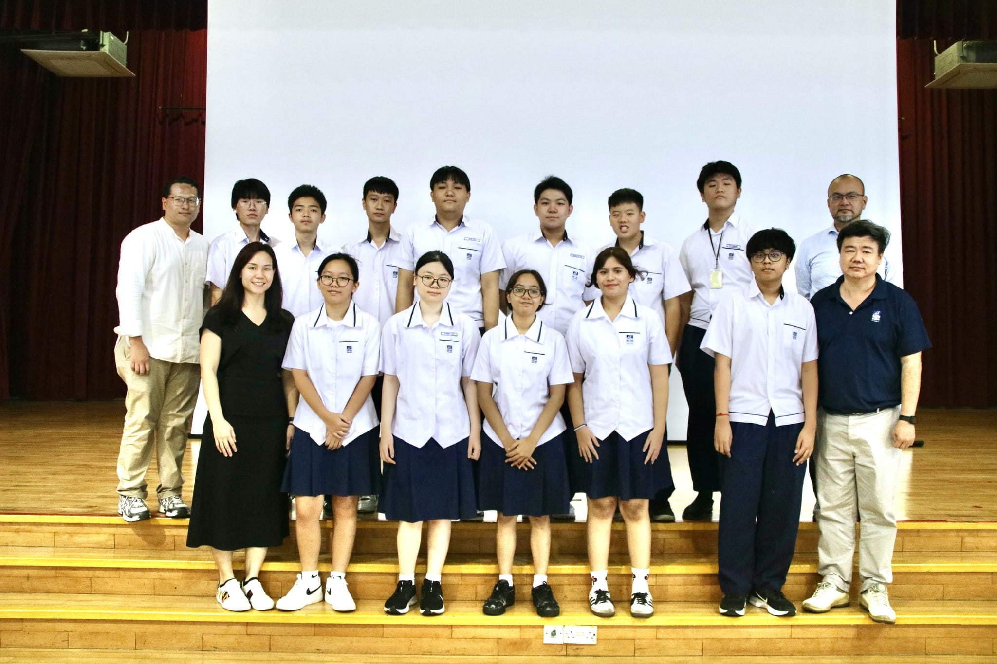 A group of students in white uniforms and teachers pose together on a wooden stage in front of a white screen.