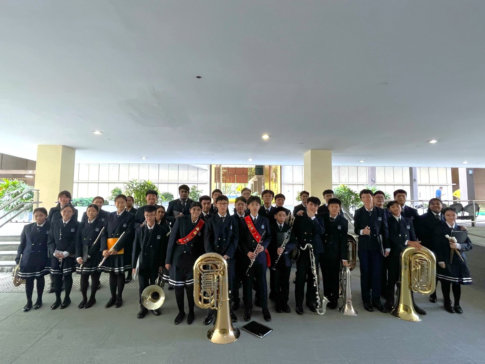 A large group of school students in dark uniforms pose together holding brass and woodwind instruments indoors.