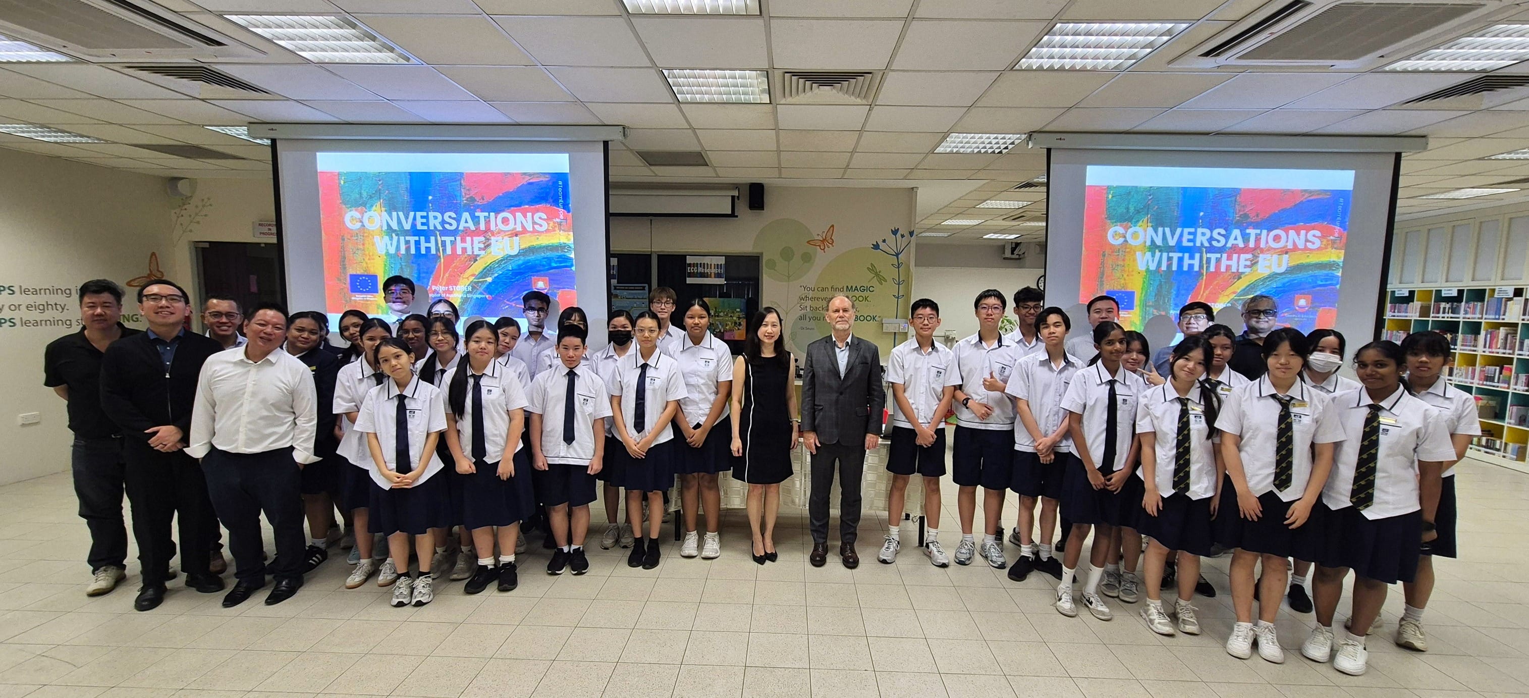 A large group of uniformed students and adults pose indoors before screens showing "Conversations with the EU."