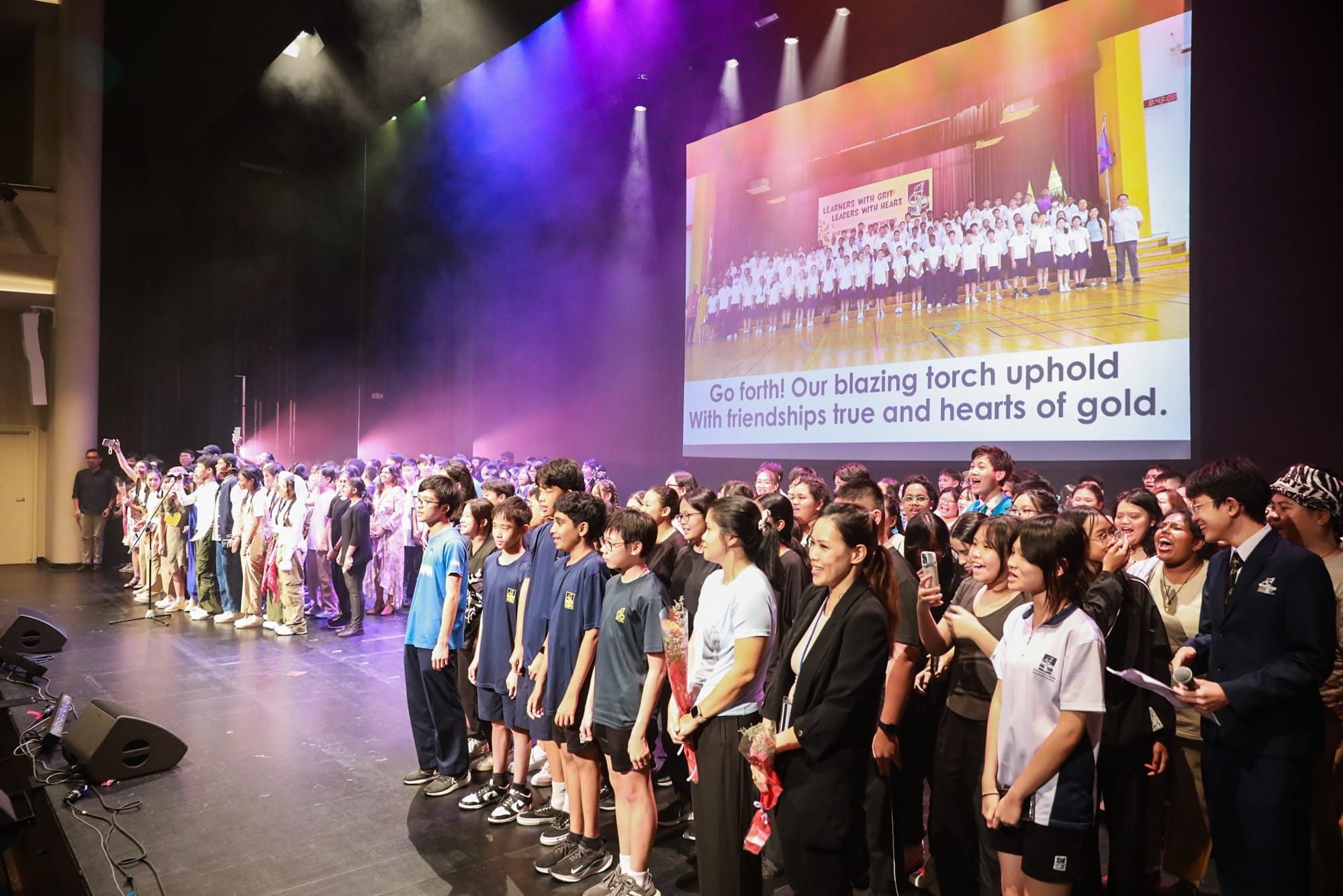Large group of students and adults on a lit stage with a projected screen showing a school song lyric.