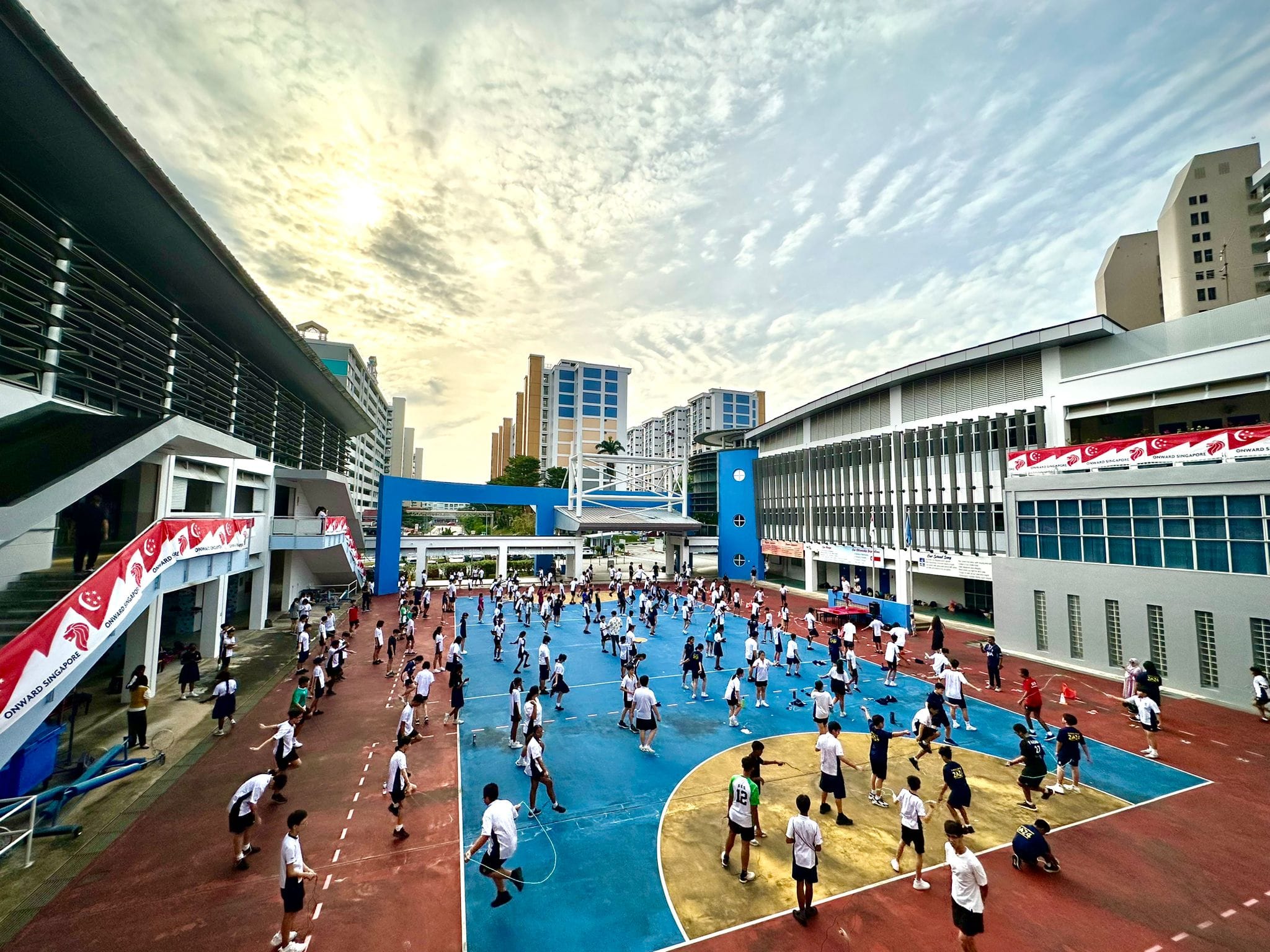 Students in white uniforms play and skip rope on a blue outdoor court between school buildings under a cloudy sky.
