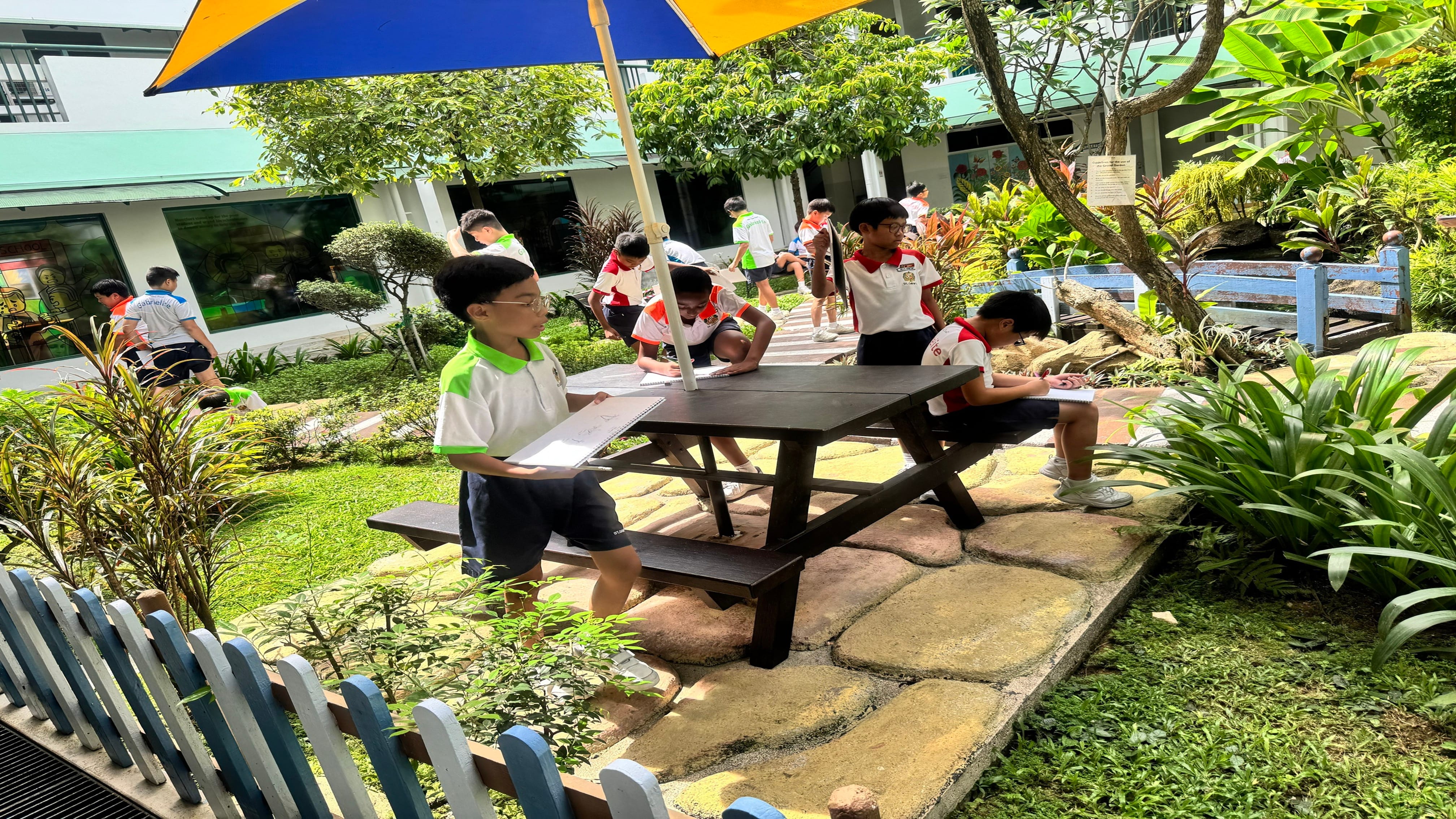 Schoolchildren draw and write outdoors around a picnic table under a large umbrella in a garden courtyard