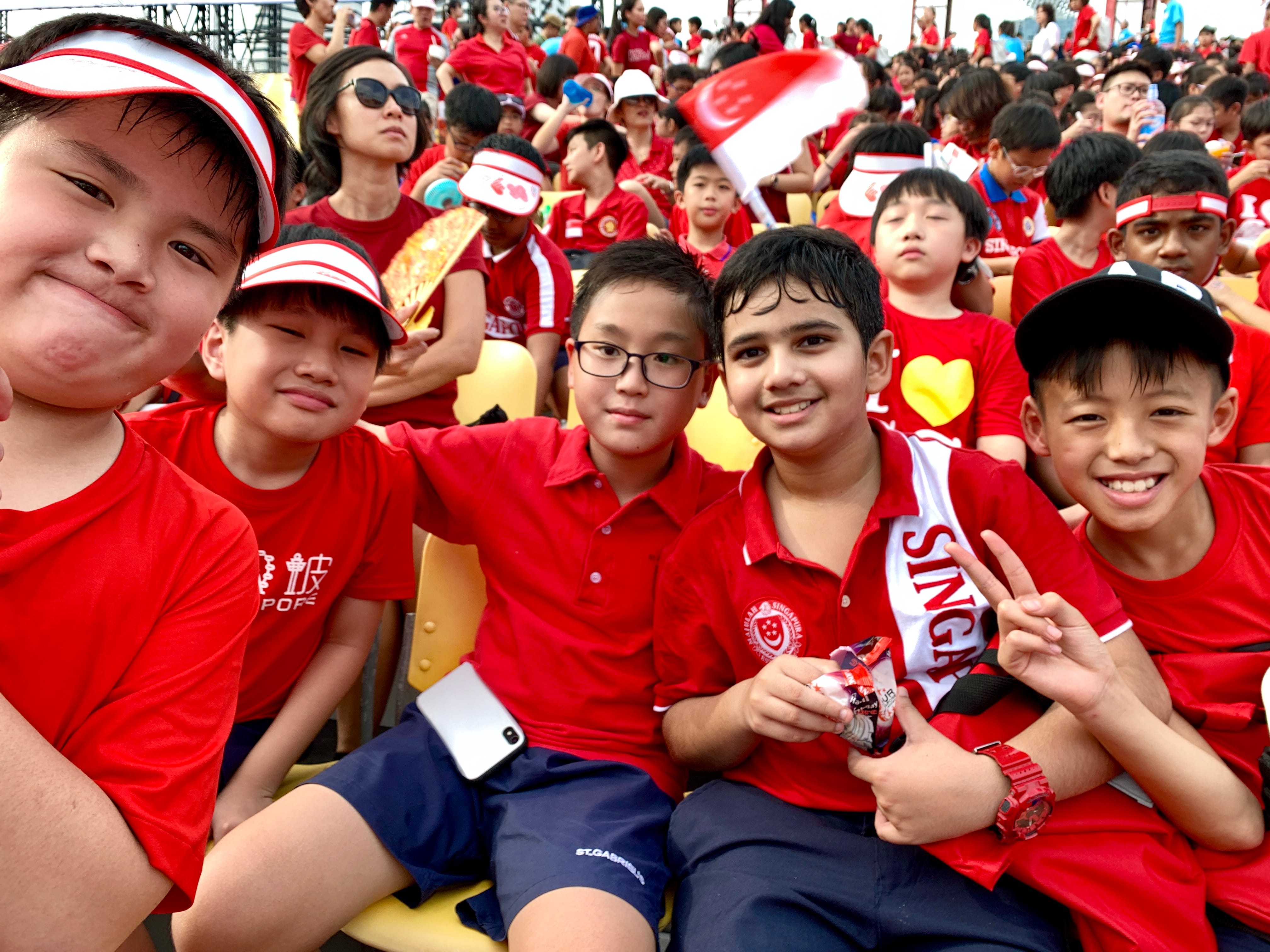 Five young boys in red Singapore shirts smile at a crowded outdoor event with a Singapore flag visible.