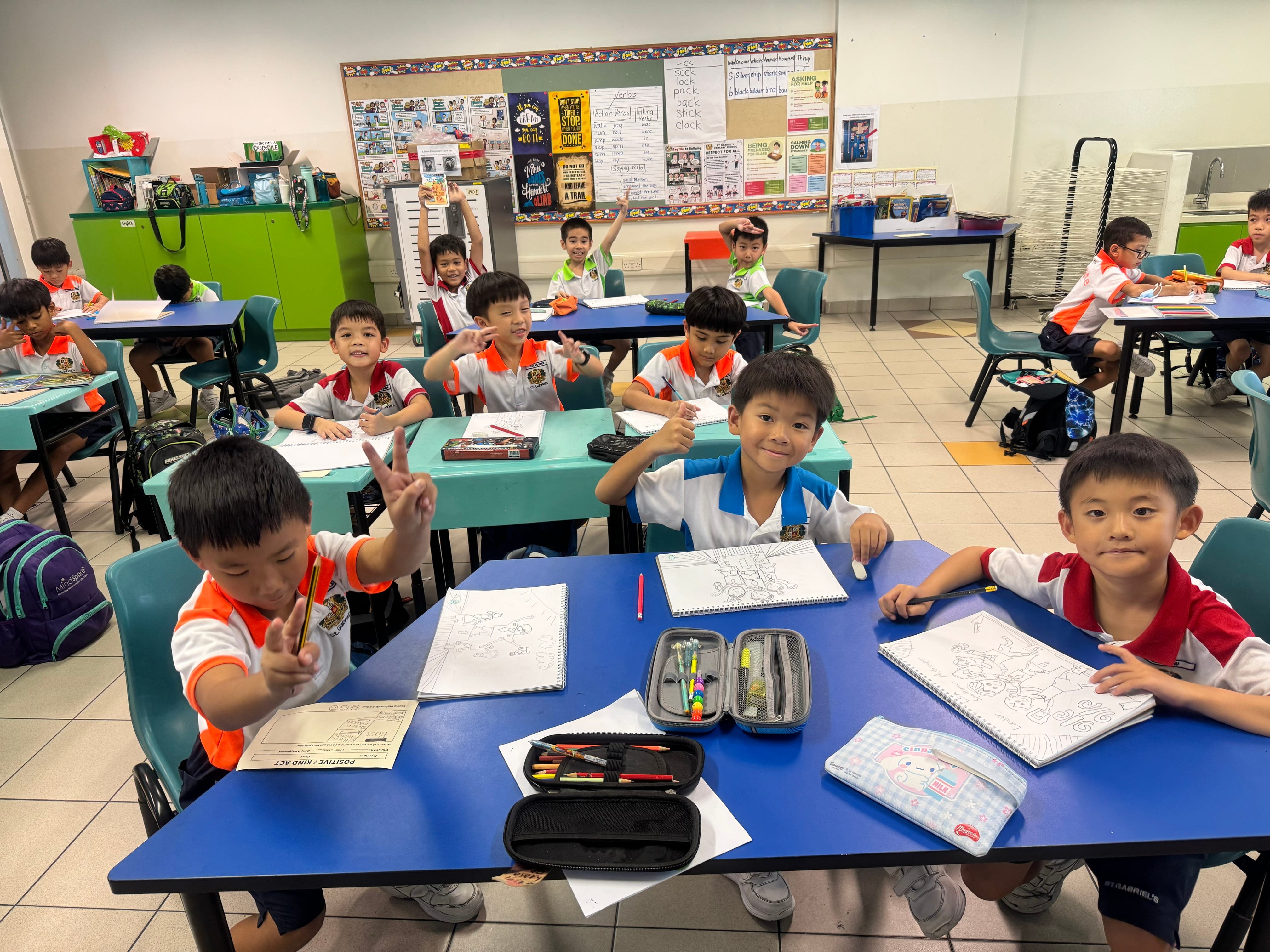 Schoolchildren at tables with notebooks and pencil cases; some smile and pose with peace signs.