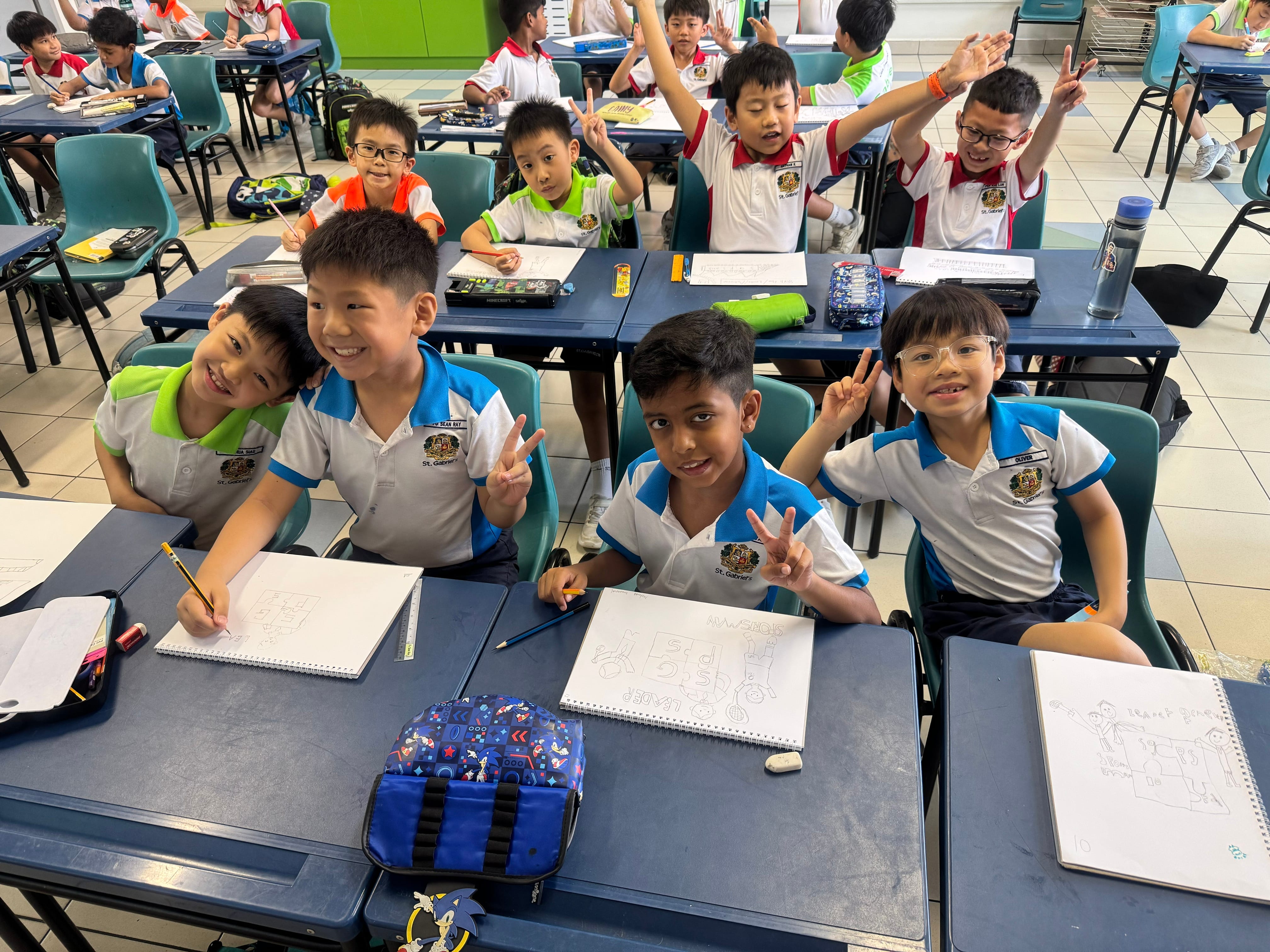 Schoolchildren at desks smiling, raising hands and flashing peace signs, with notebooks and pencil cases.