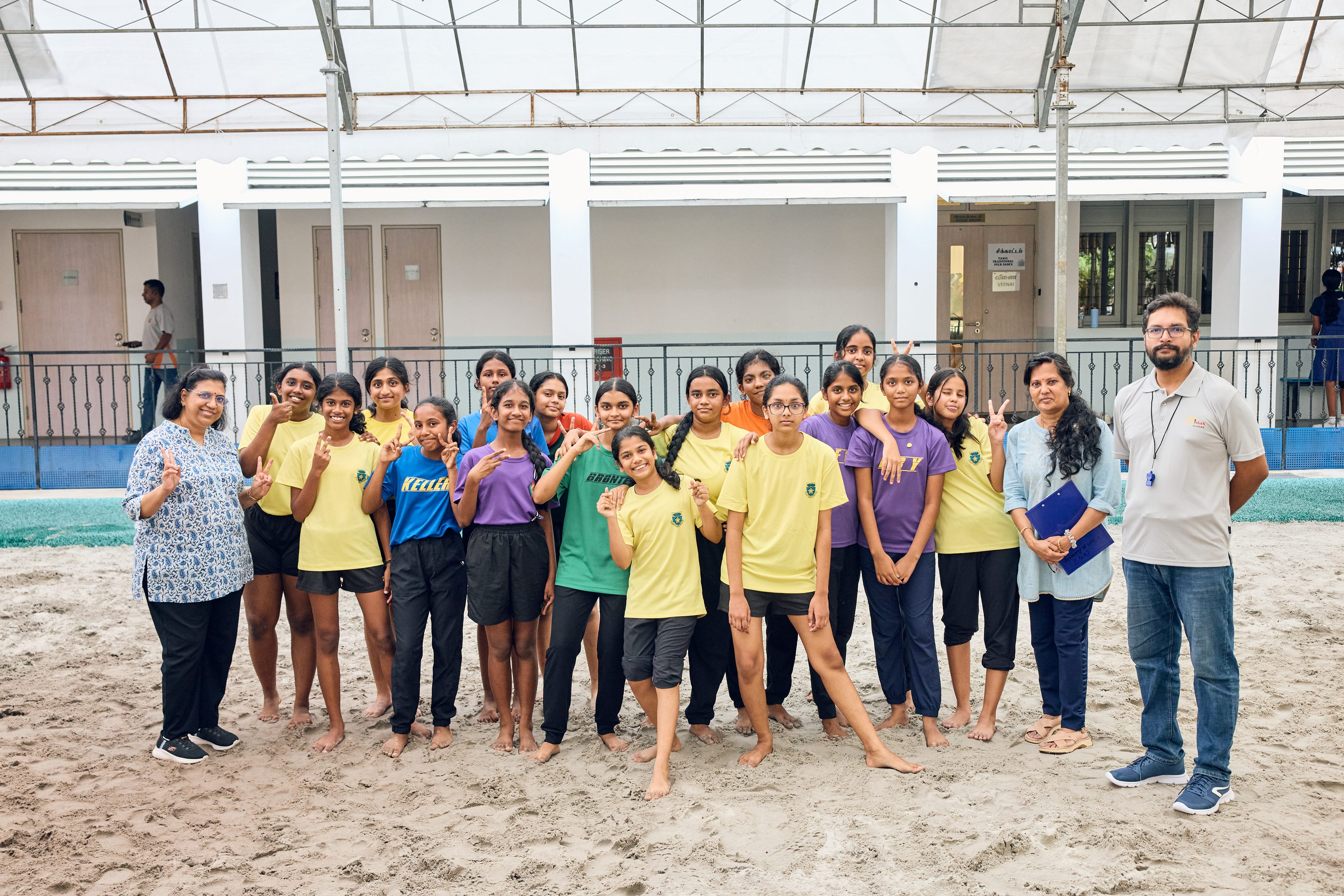 Group of barefoot girls in sports shirts pose on sand under a canopy, with two adults at the ends.