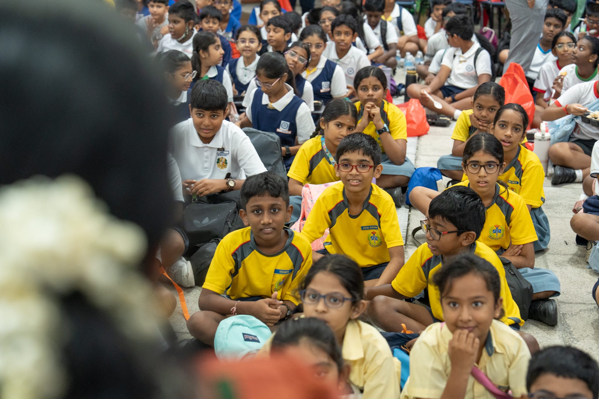 Students in uniforms sit closely on the floor, many in yellow shirts, looking toward a speaker in foreground.