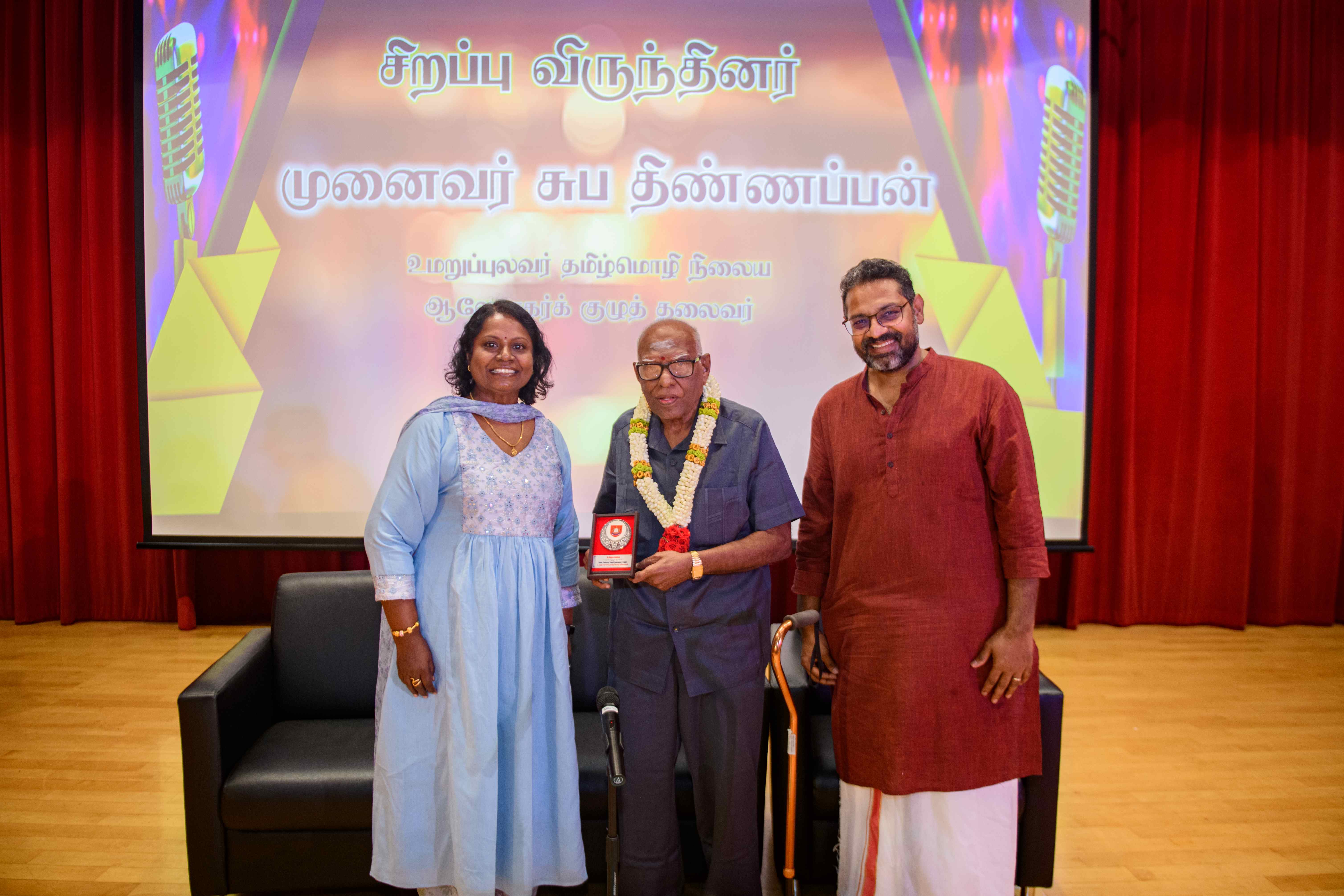 Three people pose on a stage; elderly man with garland holds plaque, Tamil text projected behind.