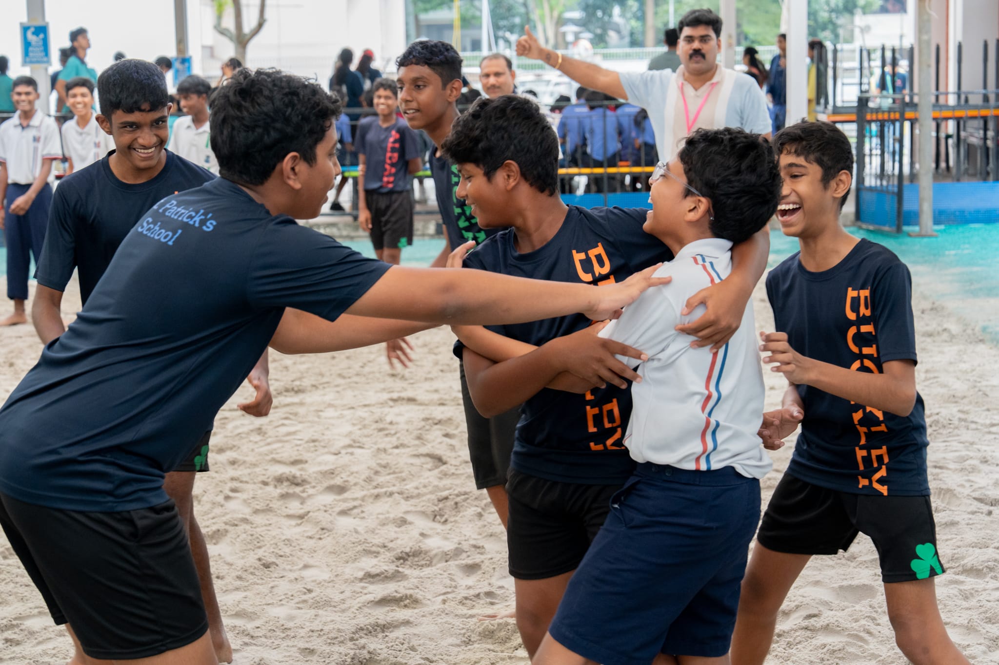 Boys laughing and hugging in celebration on a sandy court while classmates watch in the background.