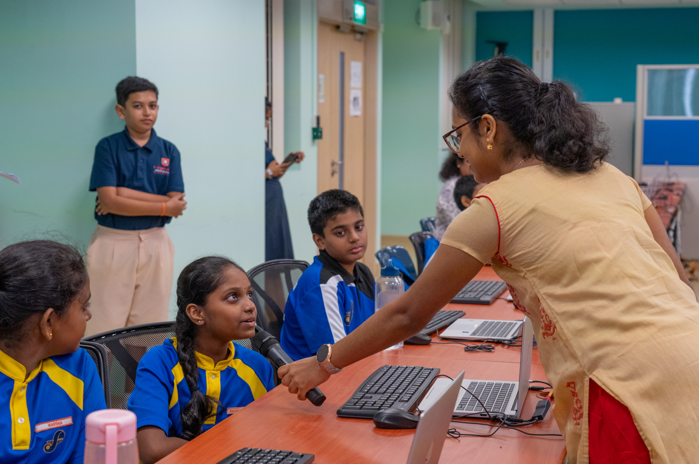 Woman holds a microphone to a seated student in a computer lab while other students watch.