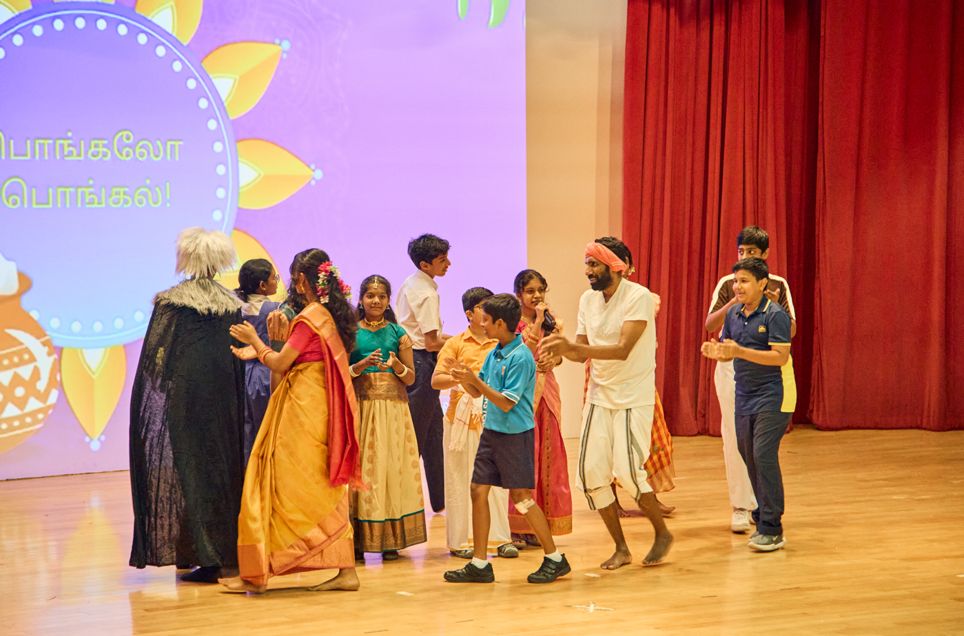 Group of children and adults on stage, clapping and acting, with Tamil text backdrop and red curtain
