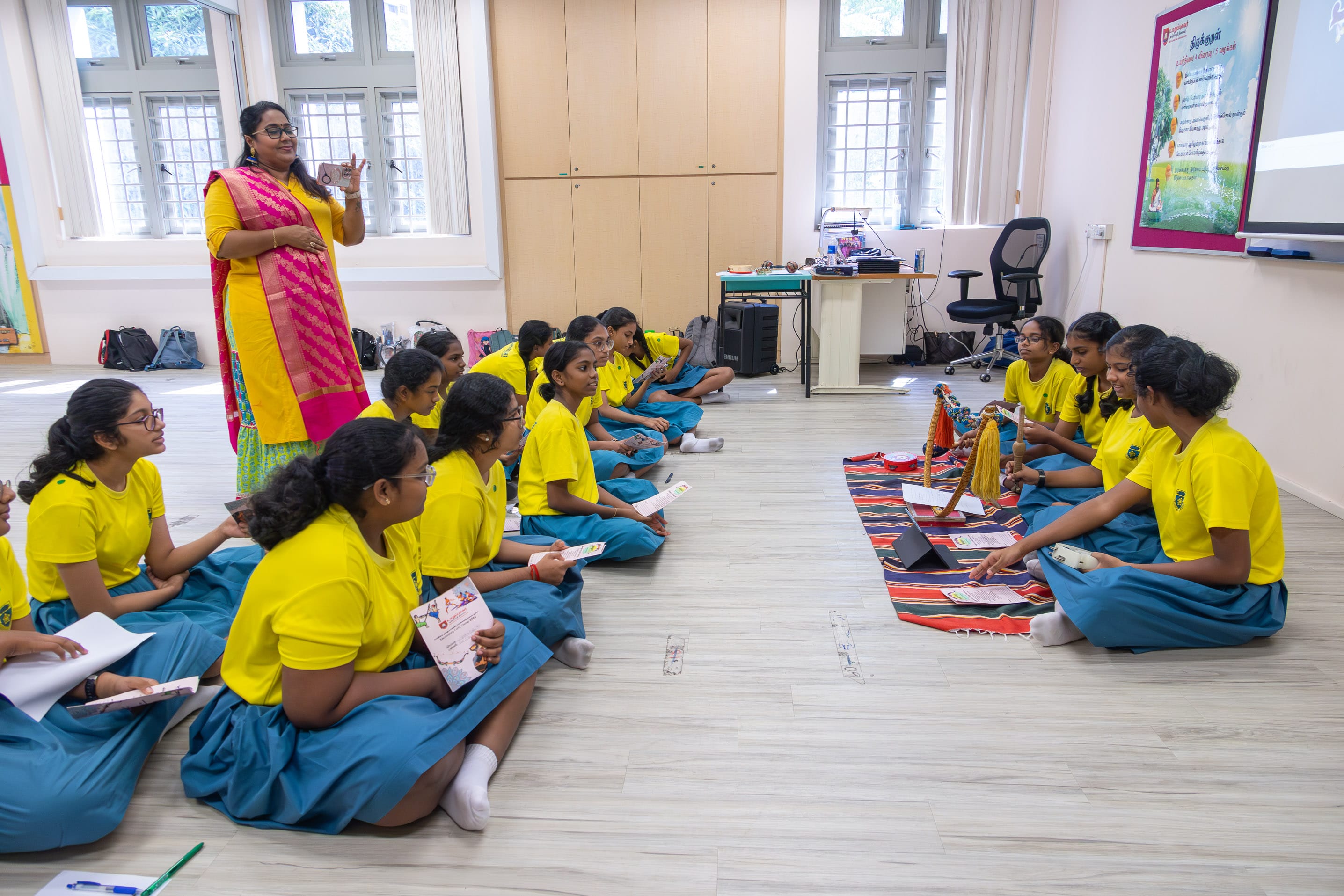 Teacher in pink and yellow stands as students in yellow uniforms sit on the floor in a classroom circle.