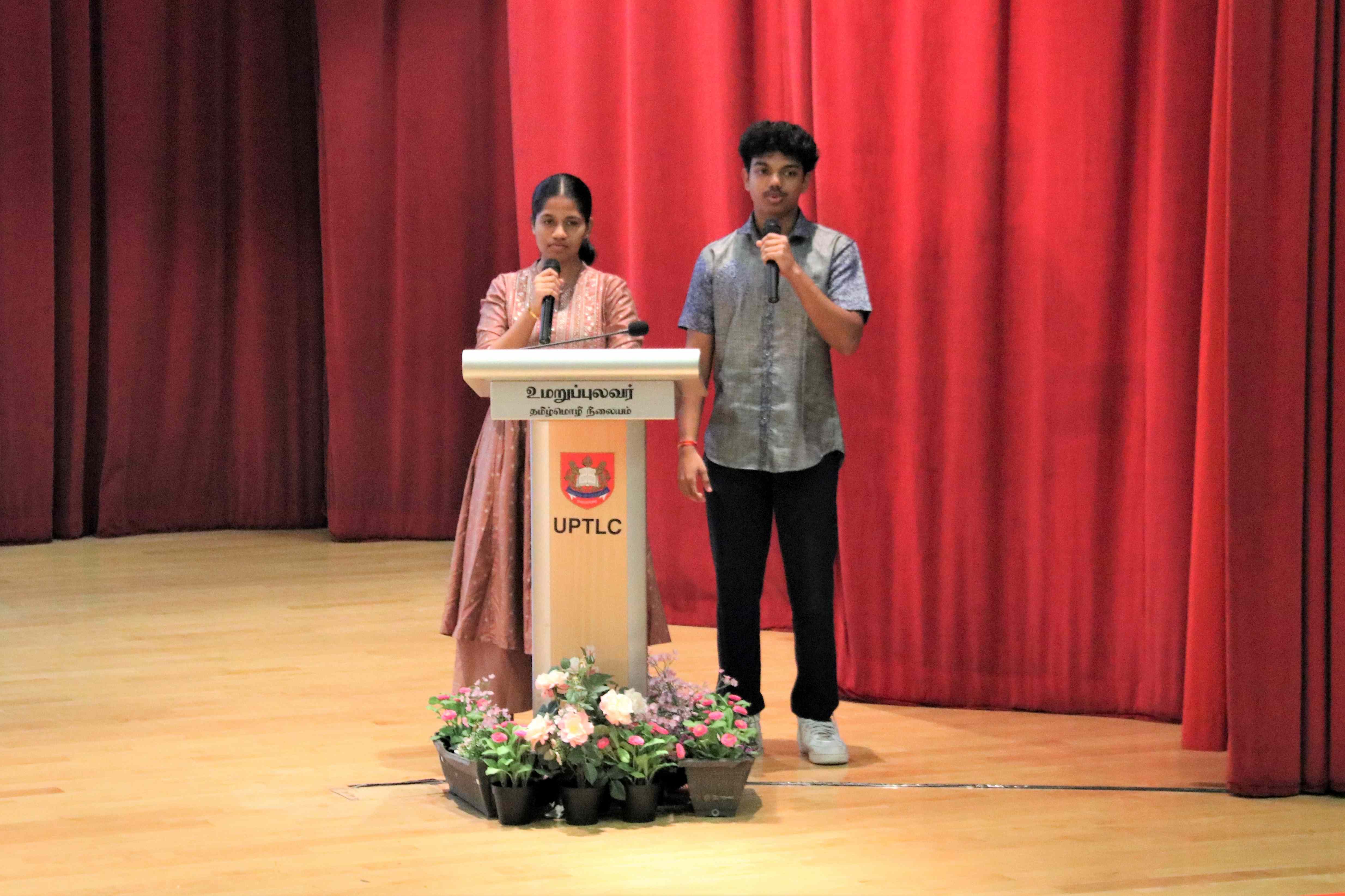 Two people on stage with microphones beside a podium labeled UPTLC, red curtains behind and flowers below.