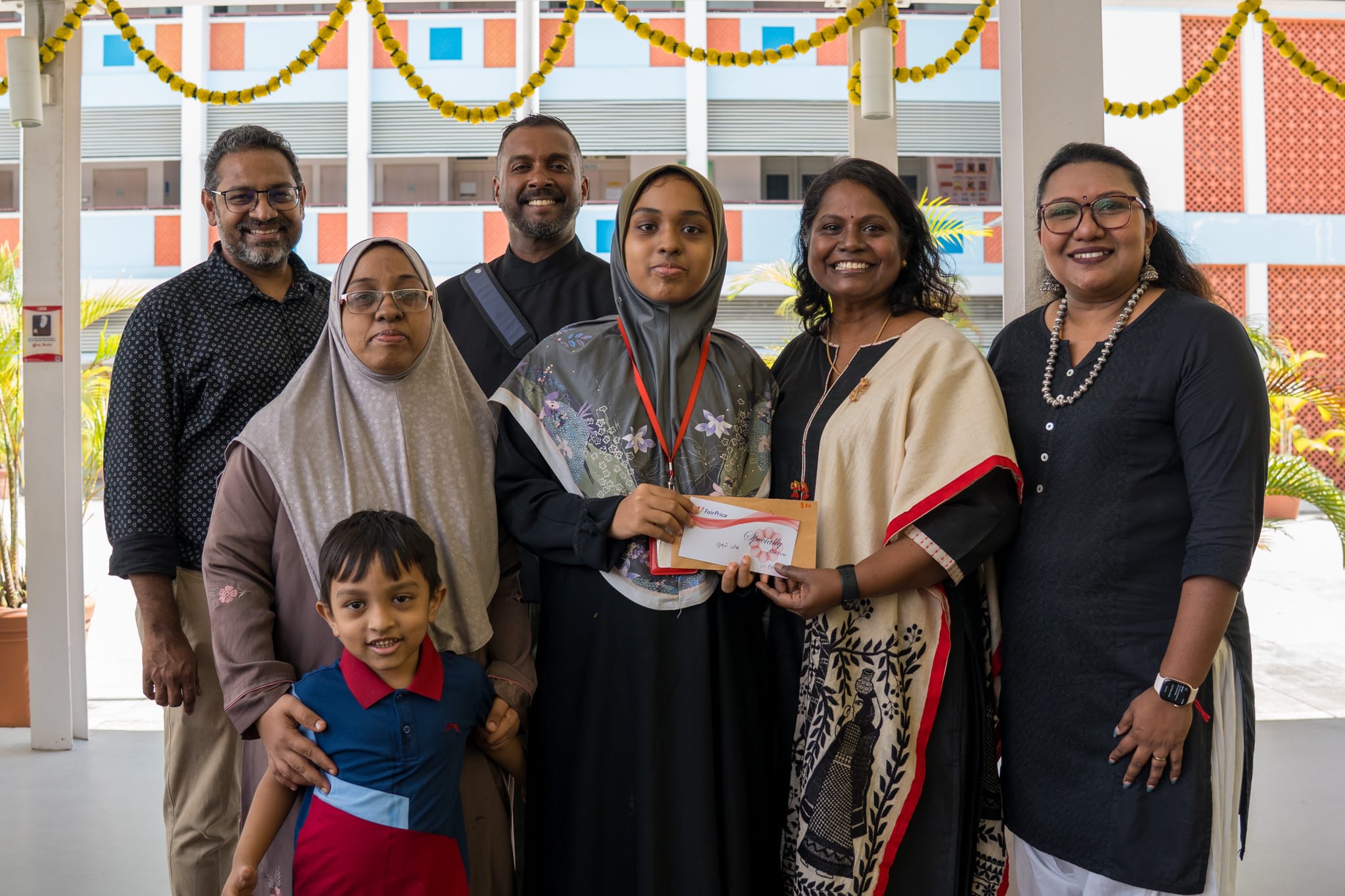 Six adults and a young boy pose indoors; two women hold an envelope, with garlands hanging above.