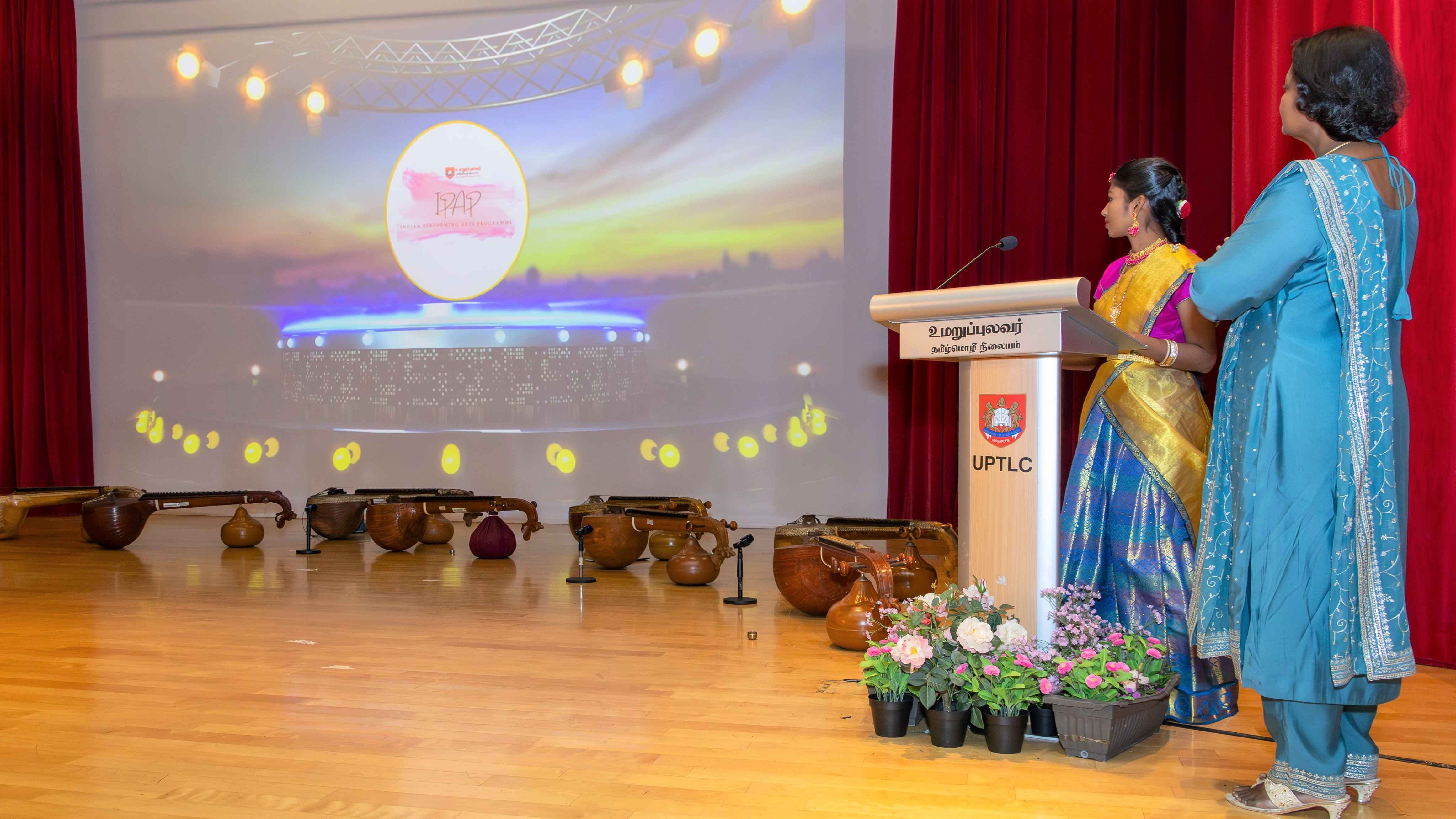 Two women at a podium on stage face a projected screen; Indian instruments lie on the floor nearby.