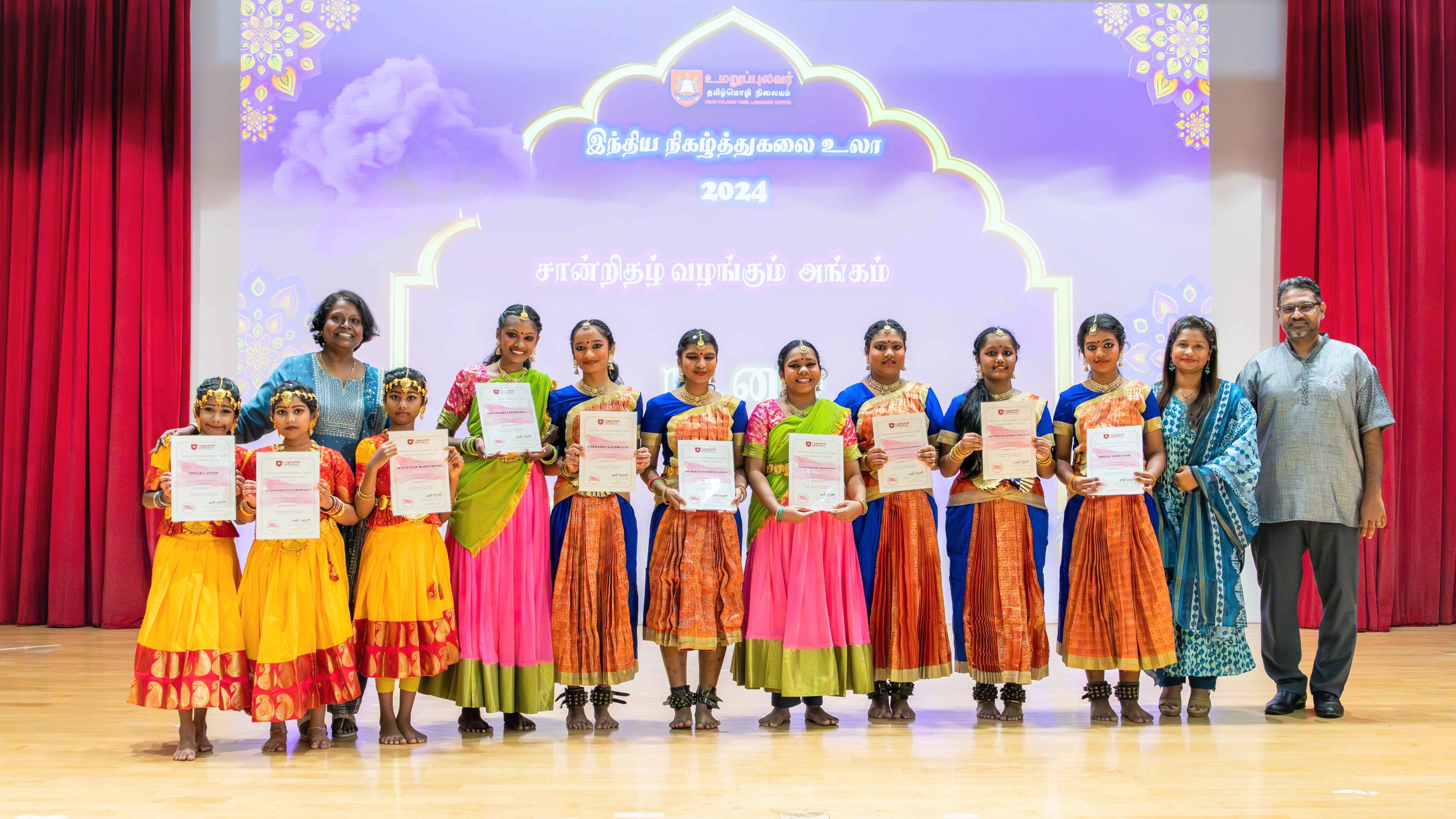Group on stage in colorful traditional outfits holding certificates, with two adults on each side.