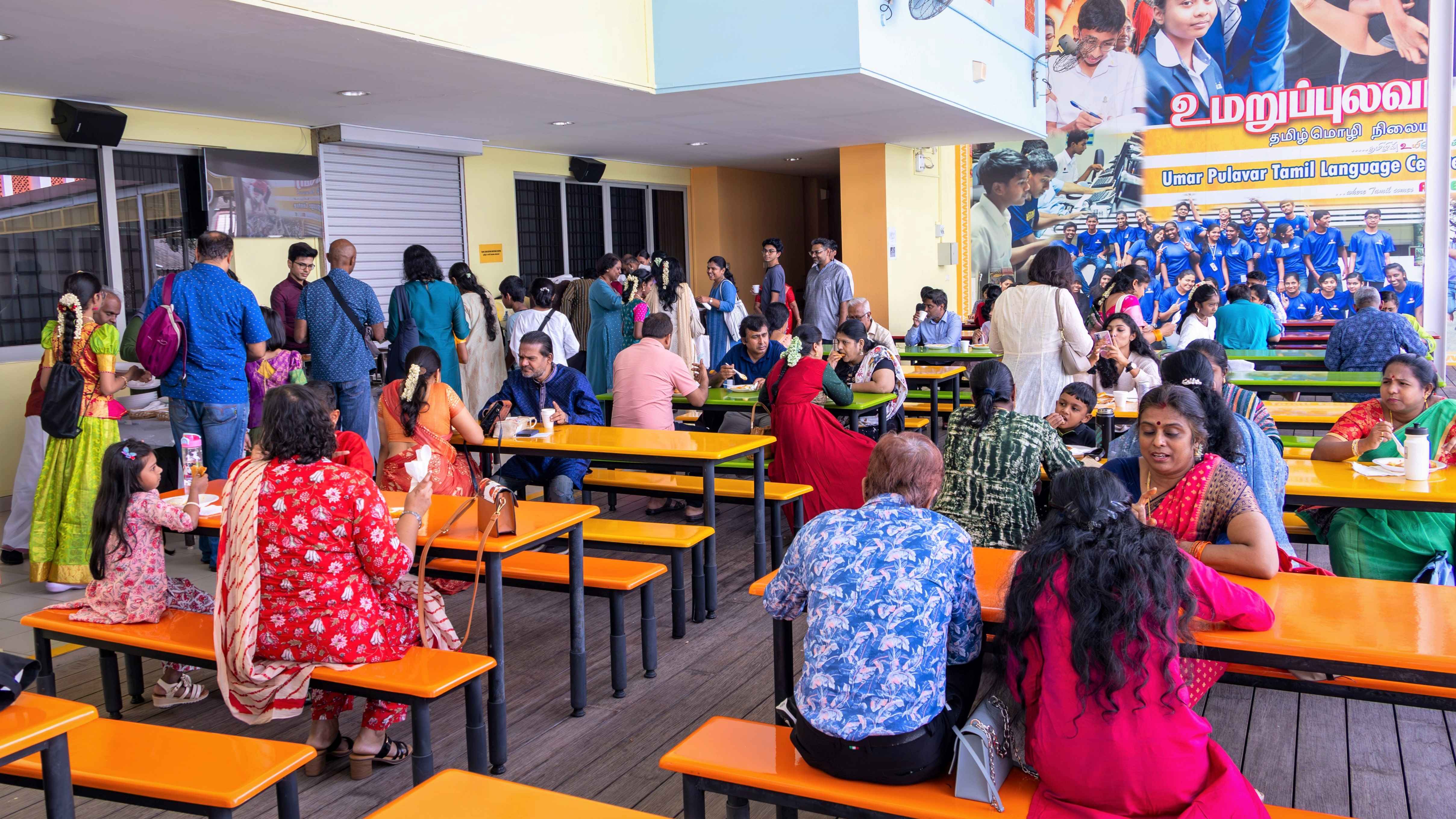 Crowded cafeteria with orange tables; people in colorful clothing eating and chatting beneath a large wall poster.