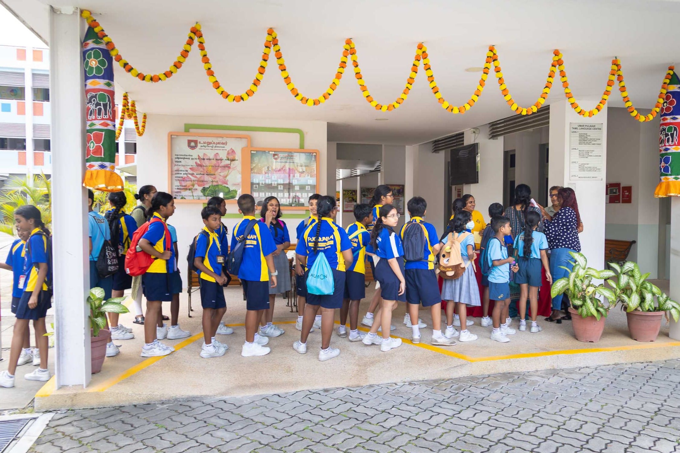 Group of schoolchildren in blue and yellow uniforms gather under marigold garlands in a corridor.