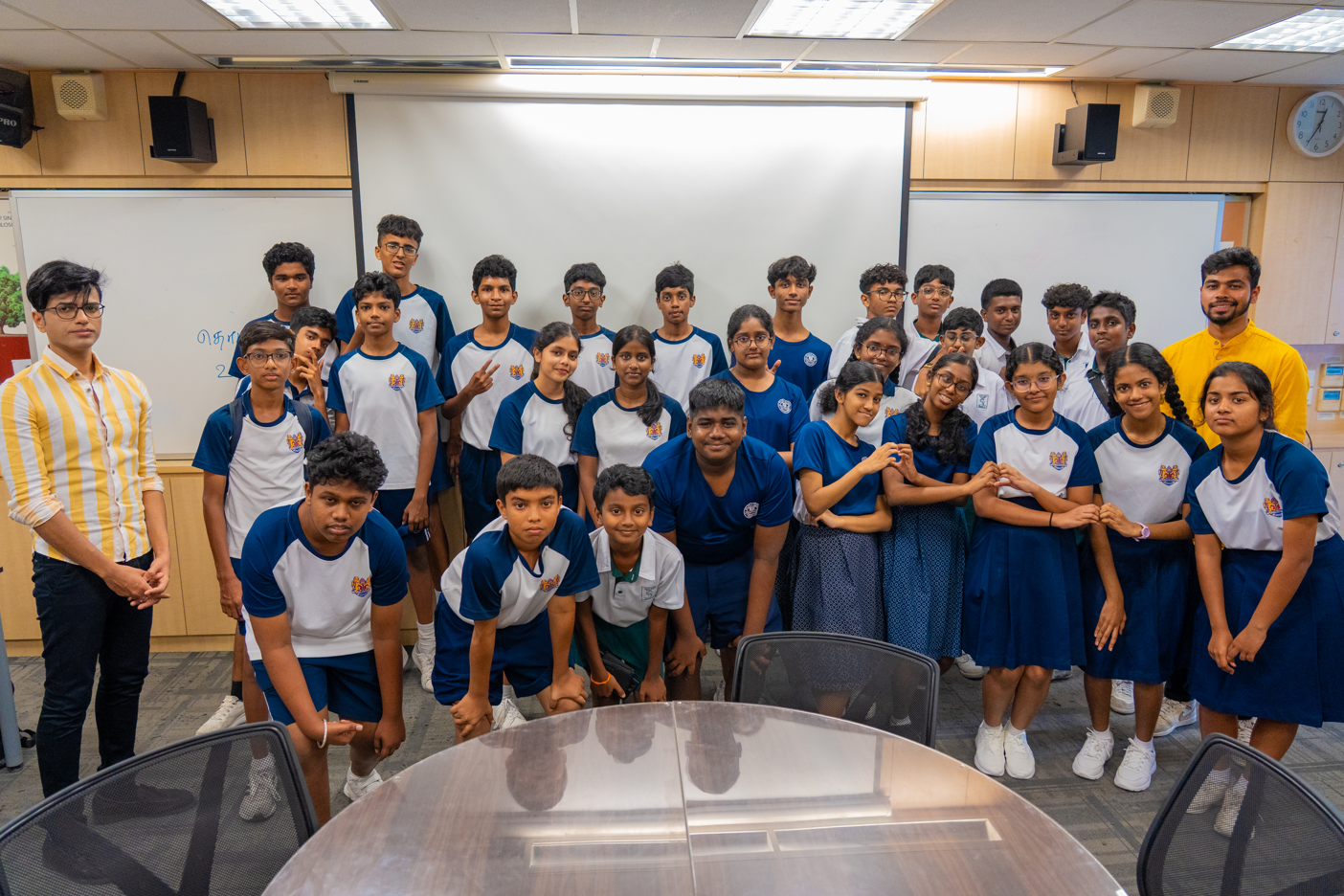 Group of students in blue and white uniforms posing in a classroom, two adults standing at sides.
