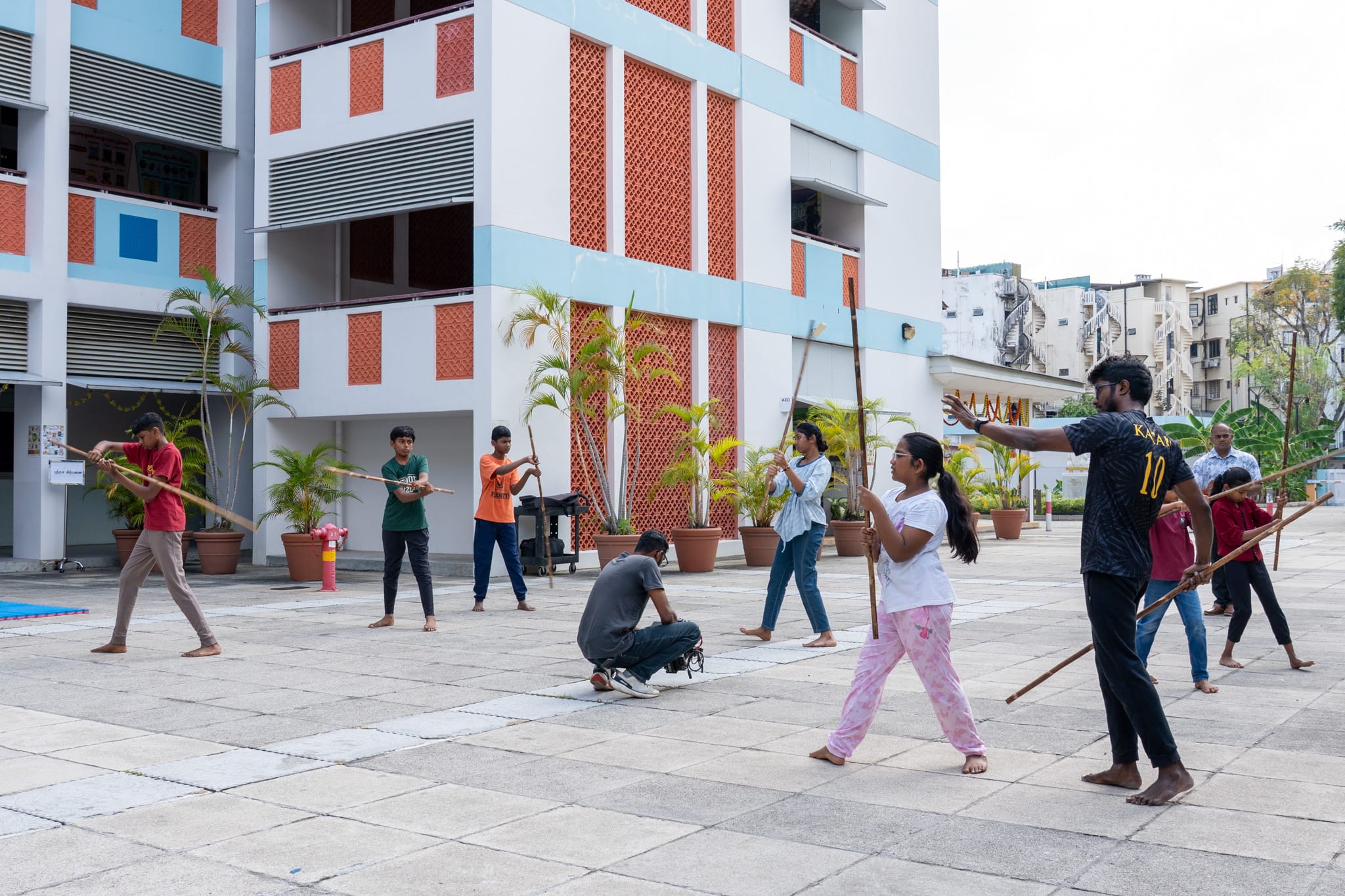 Group practices stick fighting in a courtyard outside a modern building with potted palms, one person kneeling.