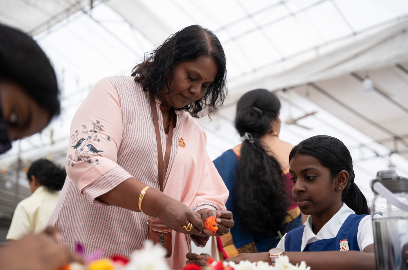 Woman in pink blouse hands an orange flower to a seated schoolgirl in blue uniform indoors.