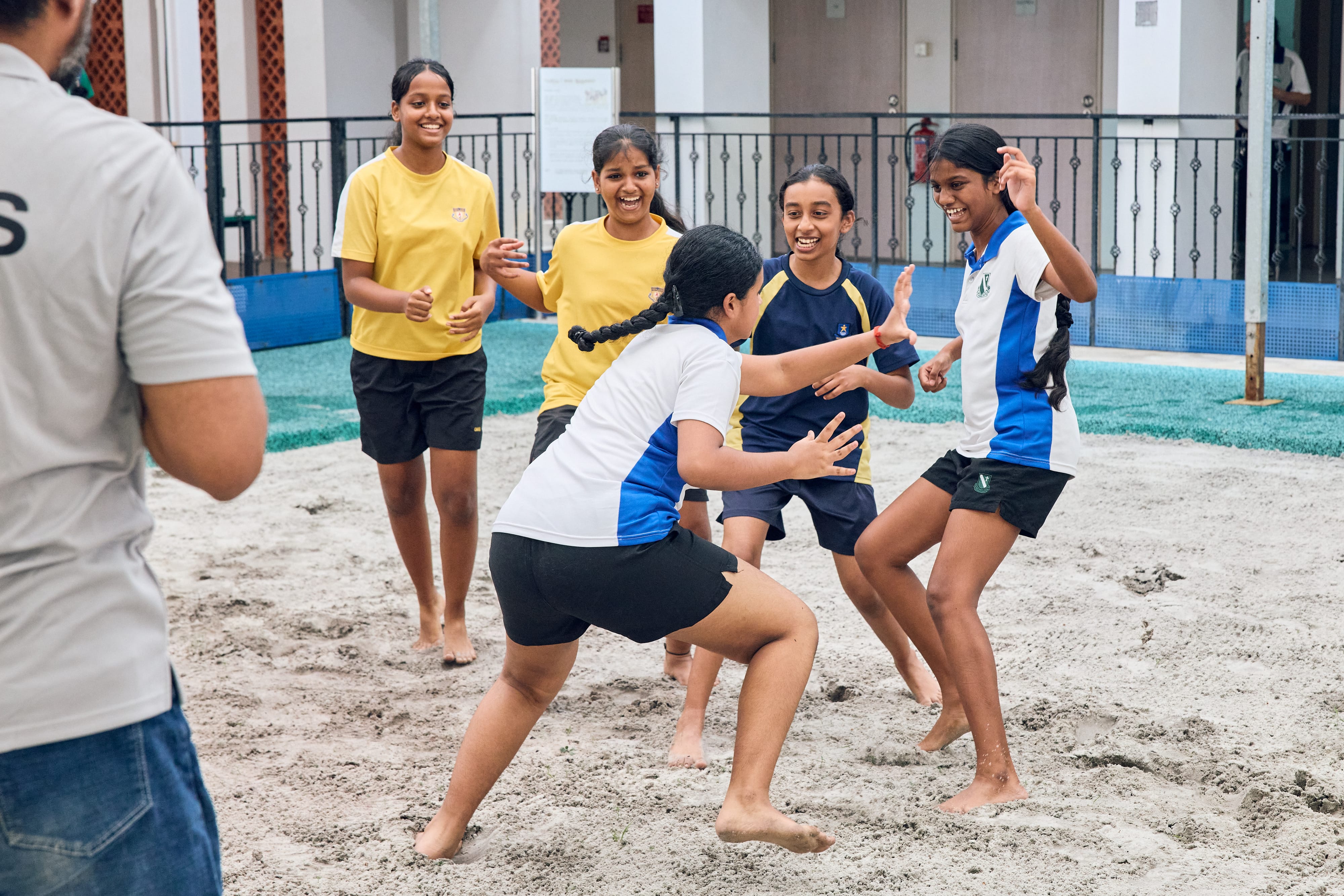 Girls in sports uniforms play on a sandy court, smiling and dodging each other while a coach watches.