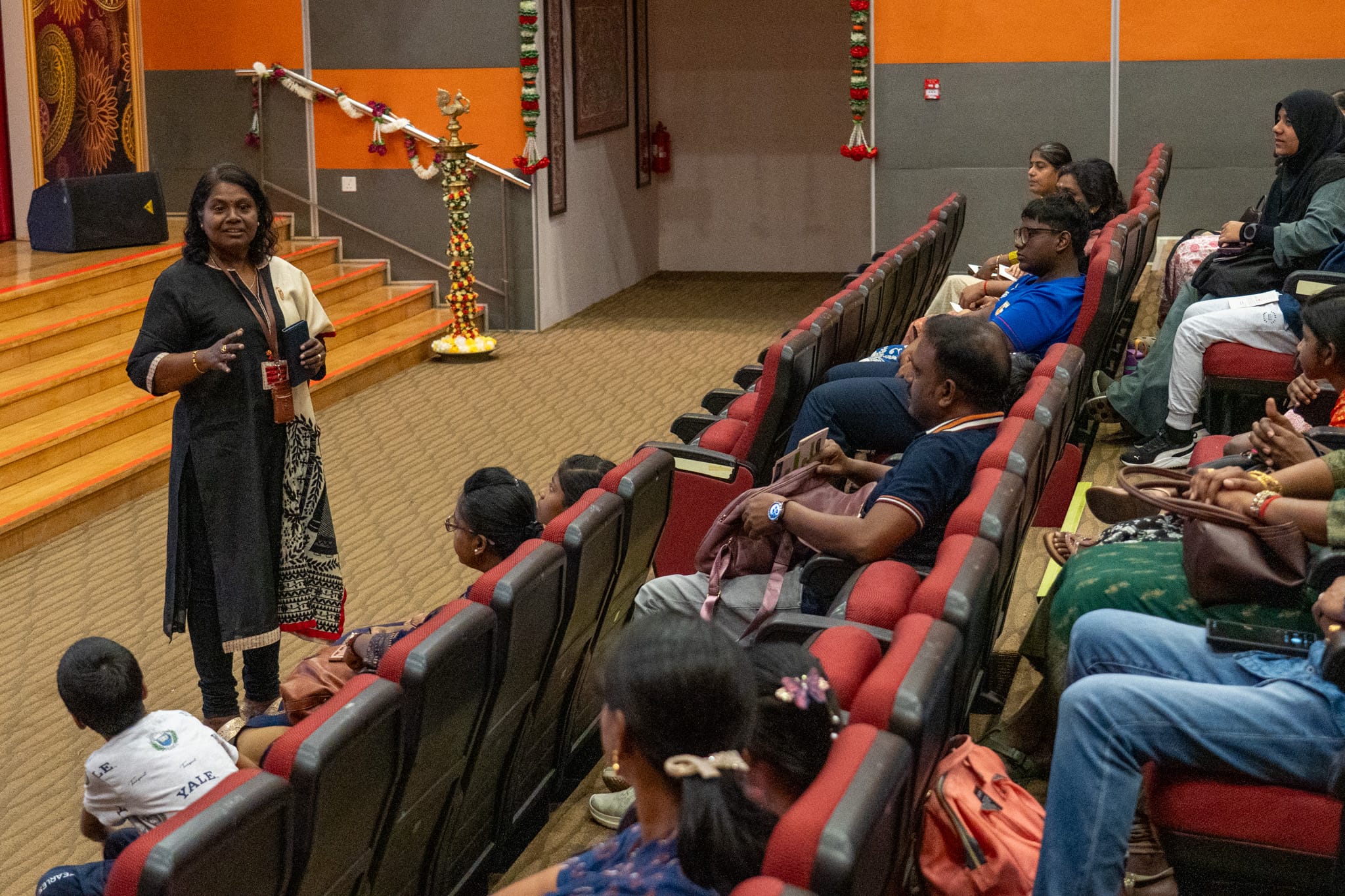 Woman speaks to seated audience in an auditorium with red chairs near a stage with steps and garlands