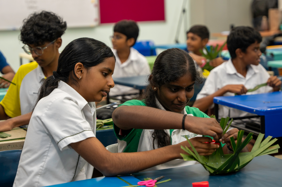Two schoolgirls at a classroom table weave green leaves, while other students work at desks behind them.